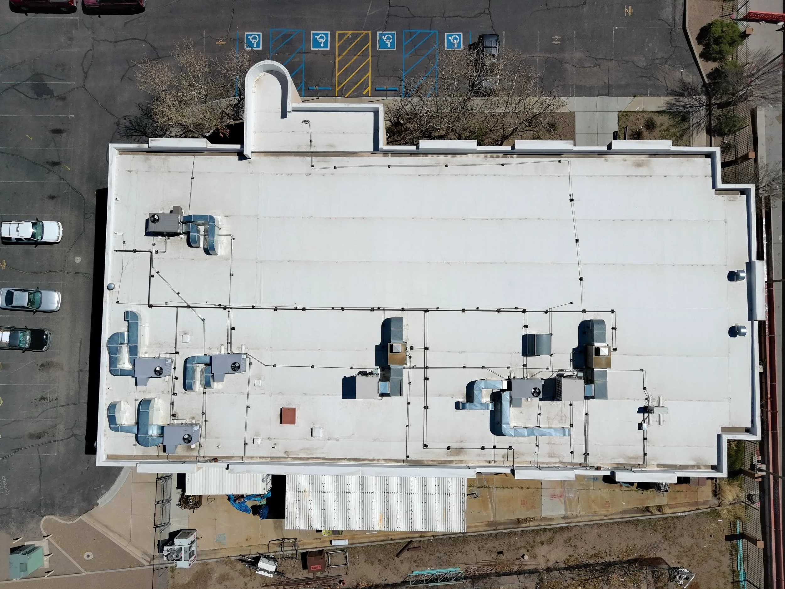 Aerial view of a rooftop with HVAC units, vents, and pipes, adjacent to a parking lot with marked handicapped spaces and a cluster of leafless trees.