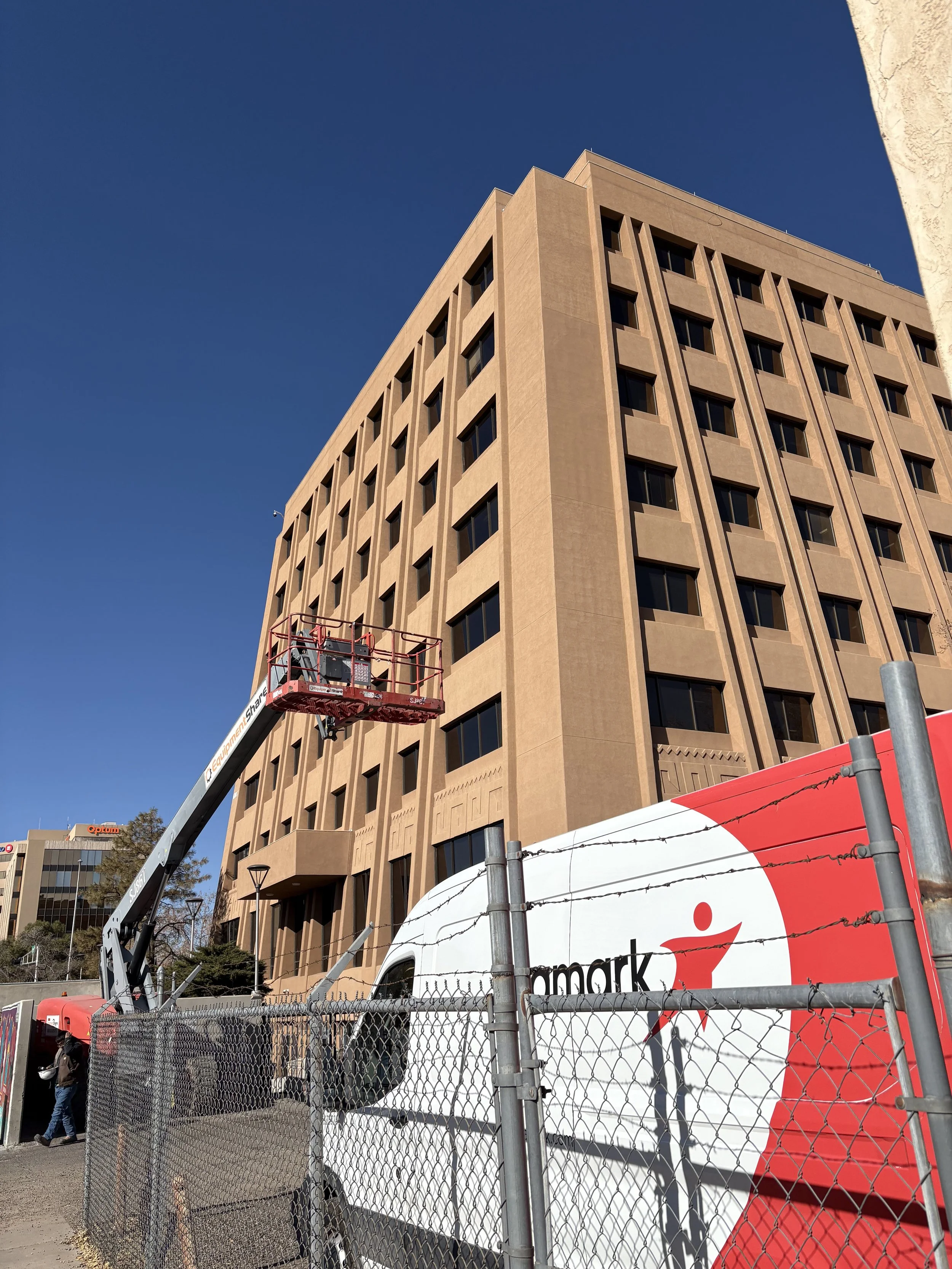A beige multi-story building under construction with a boom lift in front, a fence with barbed wire, and a sign from a retail store.