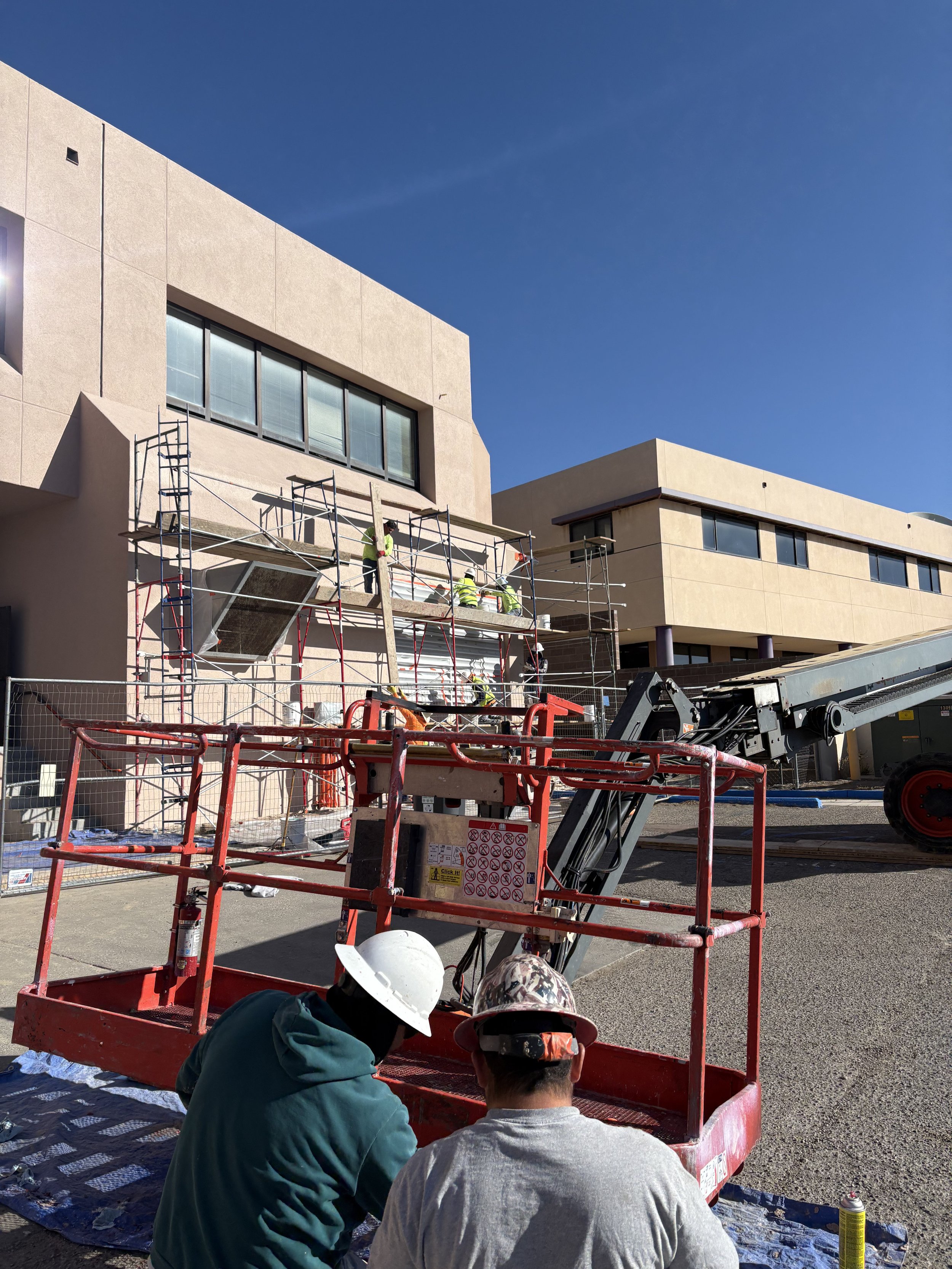 Construction workers wearing safety helmets working on scaffolding in front of modern beige buildings against a clear blue sky.