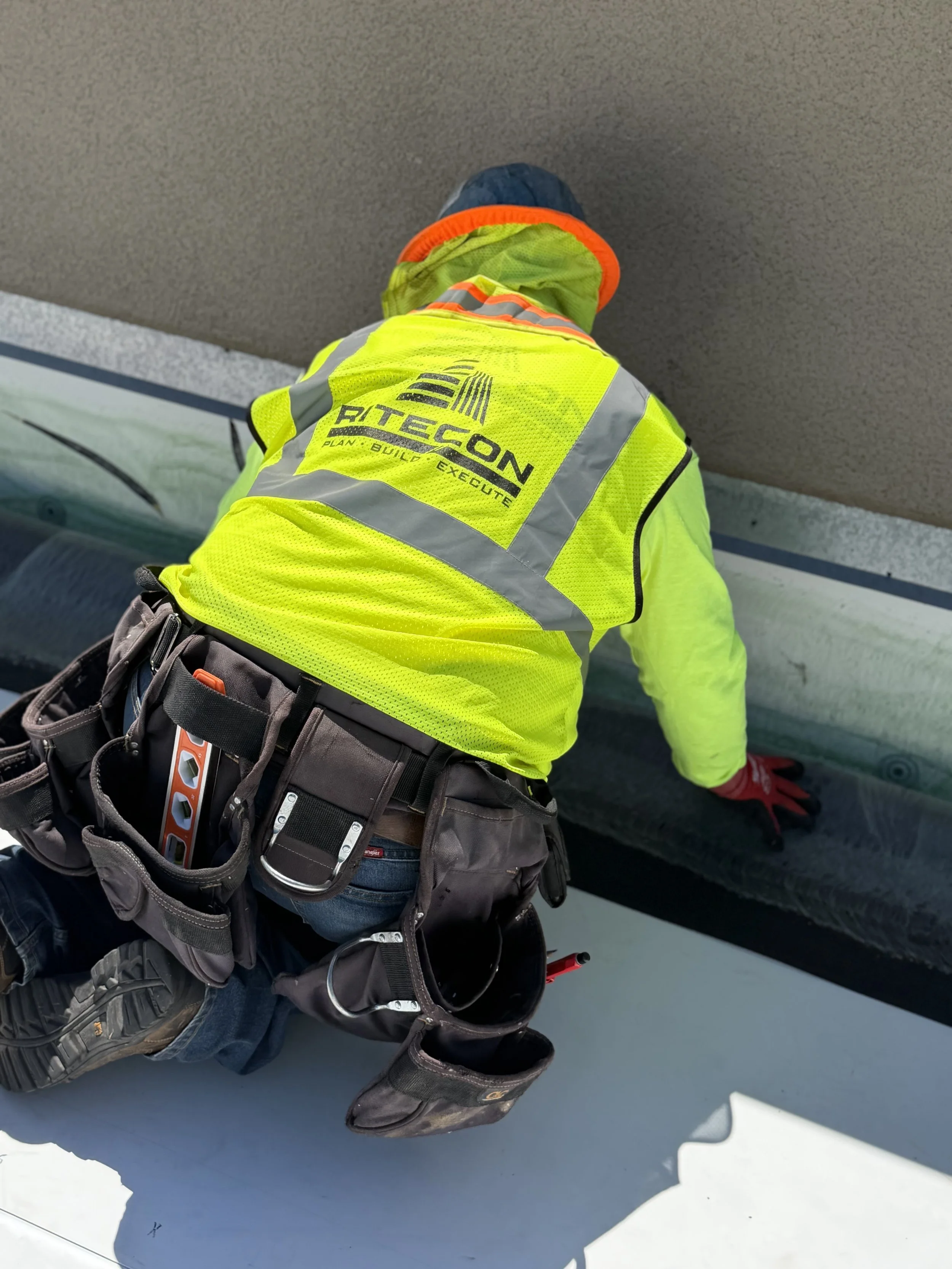 A construction worker in a yellow safety vest with the logo 'RITECON' is kneeling on the floor working on a sheet of material.