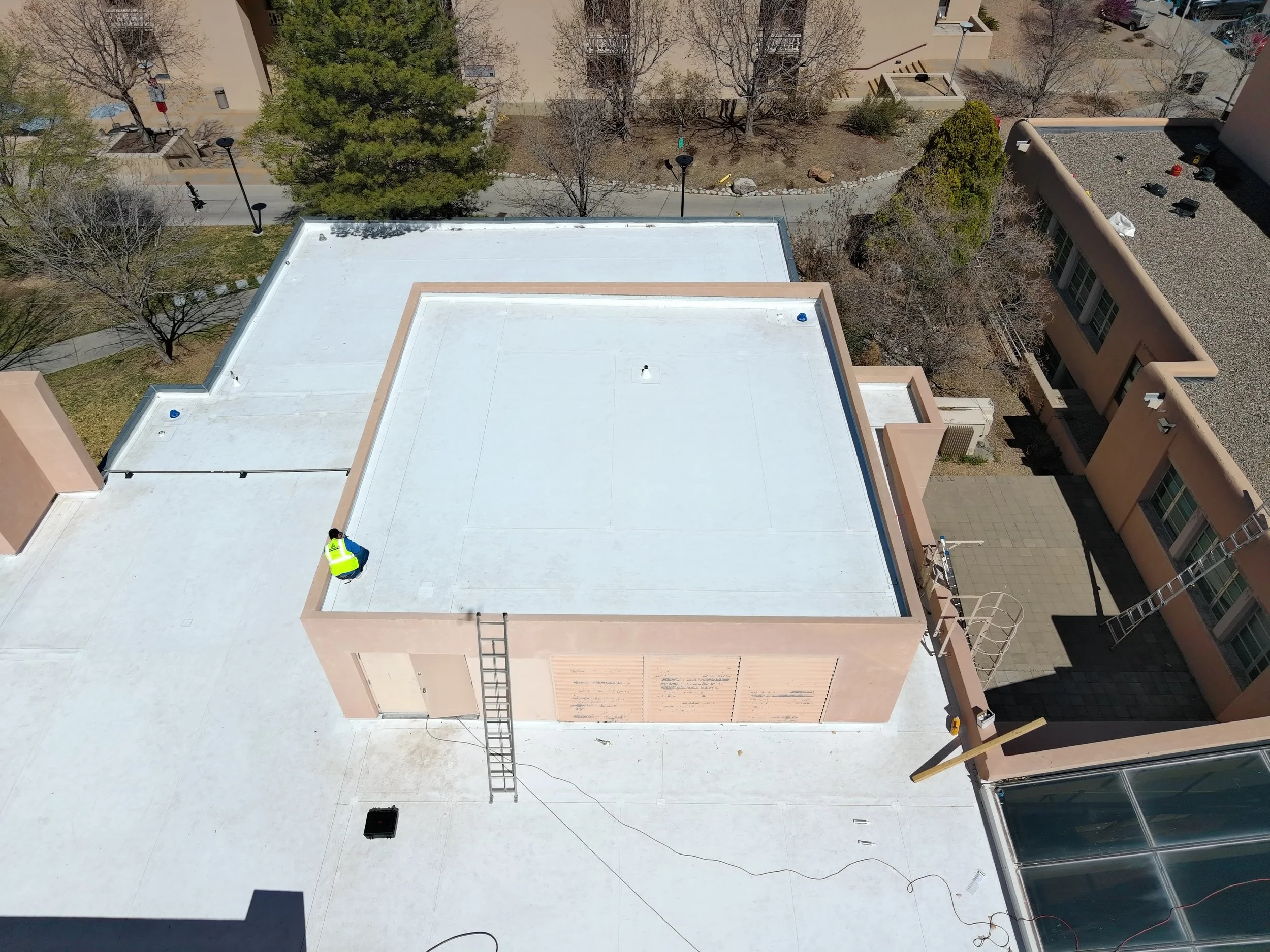 Aerial view of an outdoor flat rooftop with workers installing a white membrane, surrounded by trees, pathways, and nearby buildings.