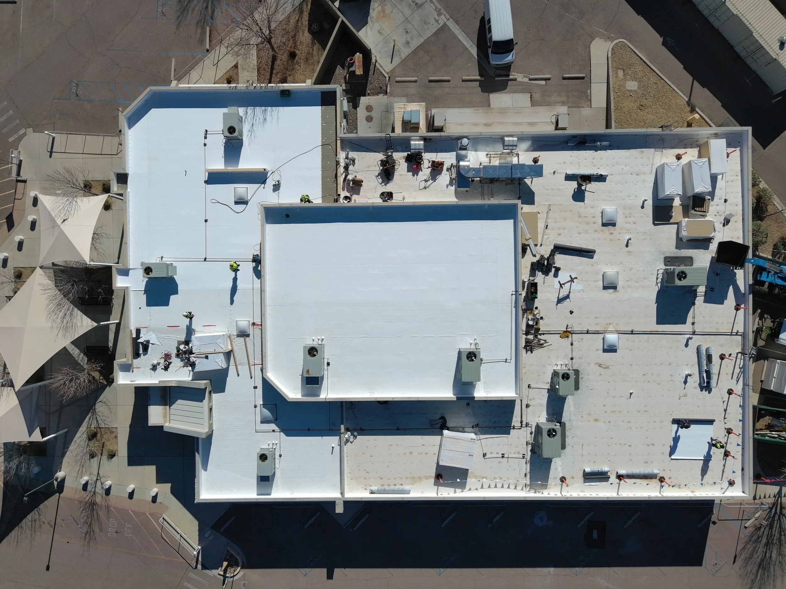 Aerial view of a building rooftop with equipment, vents, pipes, and HVAC units, surrounded by parking lot and streets.