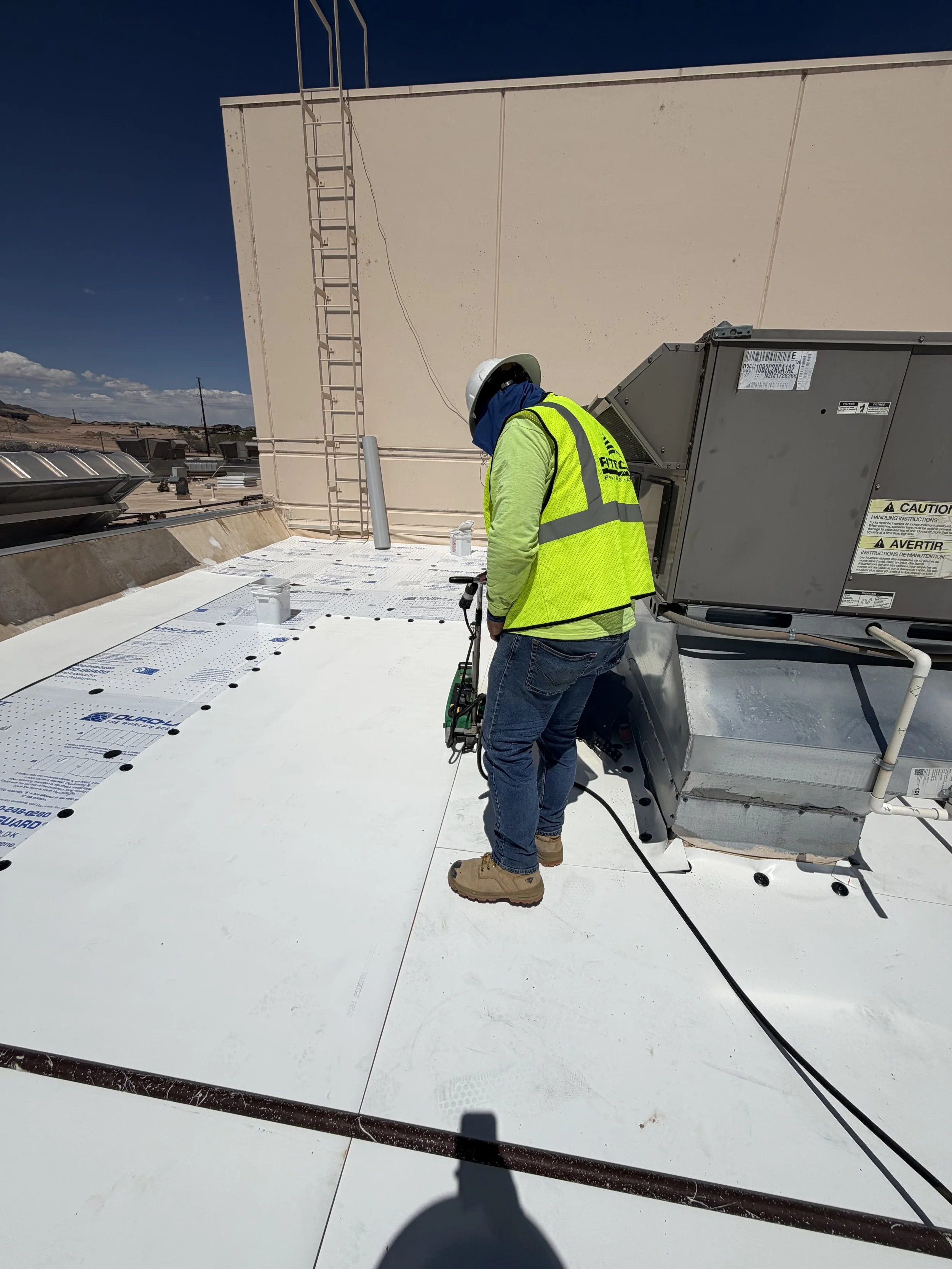 A construction worker in a neon yellow safety vest, jeans, and tan safety boots is working on a flat rooftop under a clear blue sky. He wears a white helmet and gloves, and uses equipment connected by a black cable. The rooftop has white material she