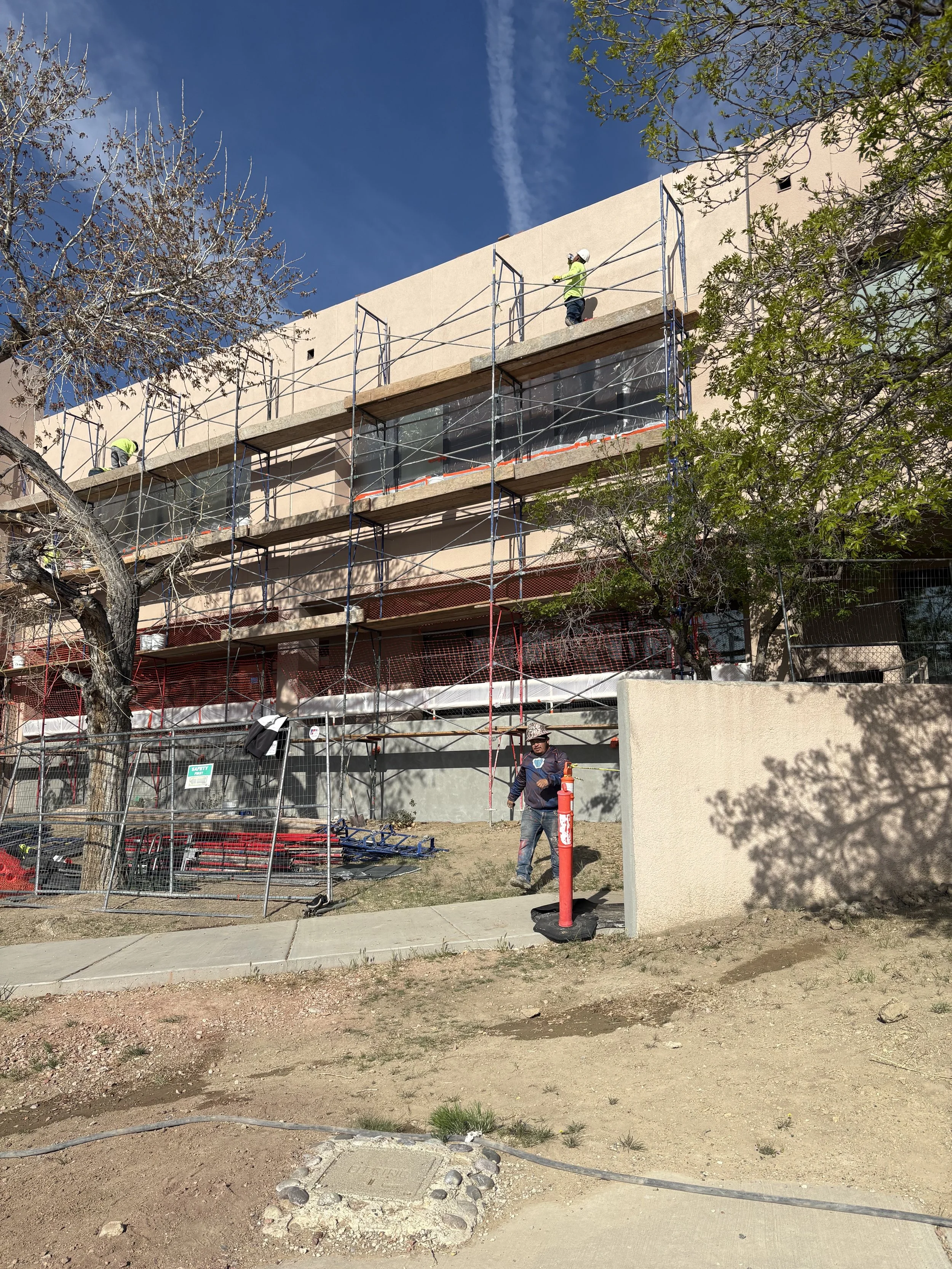 A construction site with scaffolding attached to a beige building, workers wearing safety gear on the scaffolding, trees with some leaves missing and others green, a clear blue sky, and a sidewalk and dirt in the foreground.
