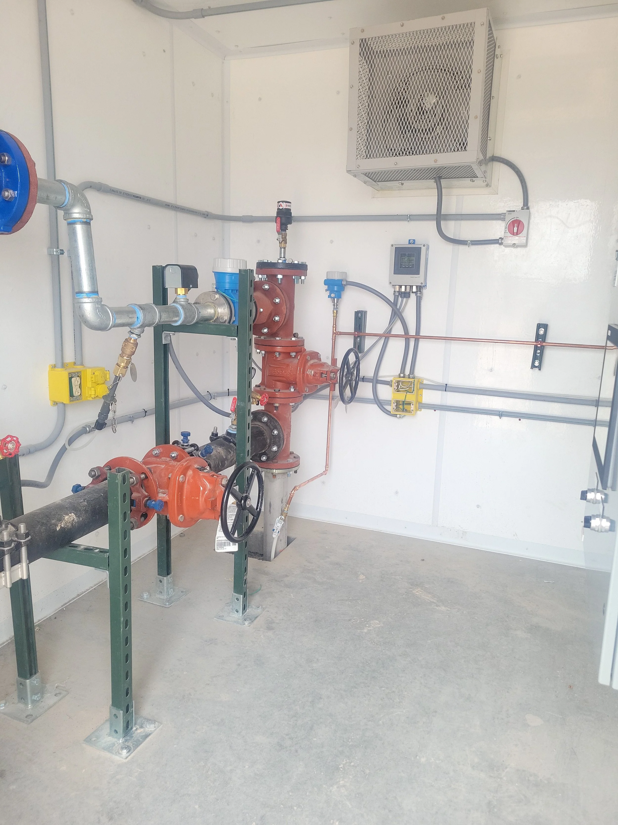 Interior of a utility or equipment room with pipes, valves, and control components mounted on metal supports and a wall. There is an air or ventilation unit mounted on the wall.