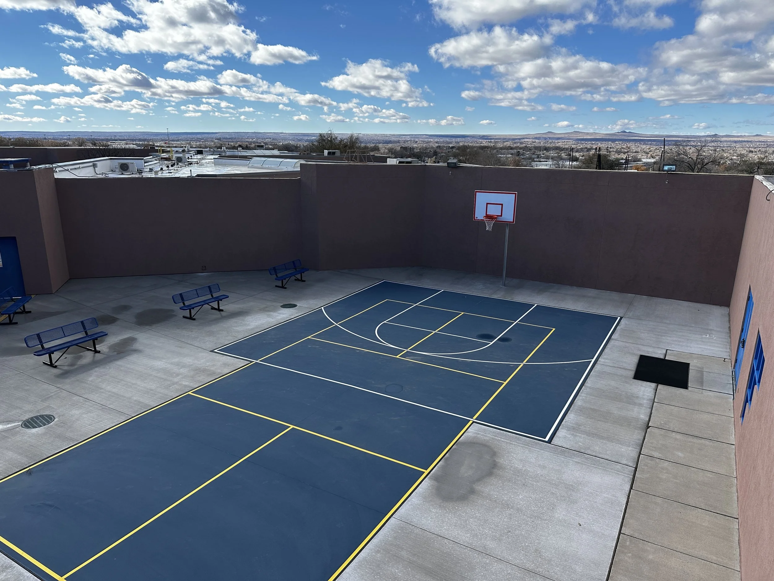 An outdoor basketball court with blue benches, a basketball hoop, and a view of a cityscape and mountains in the distance. The sky is partly cloudy.