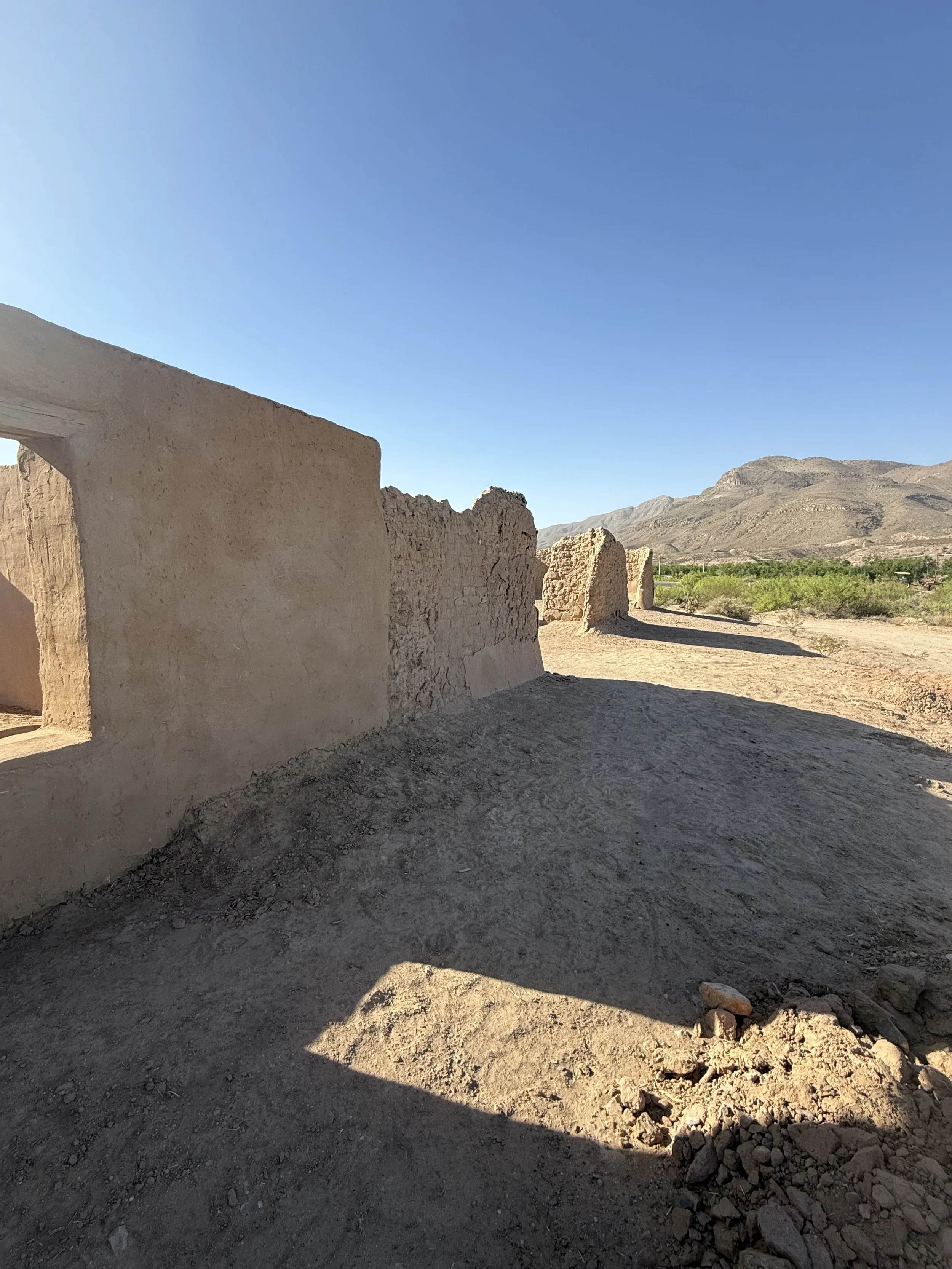 Ruins of an old adobe building in a desert landscape with mountains and a clear blue sky in the background.