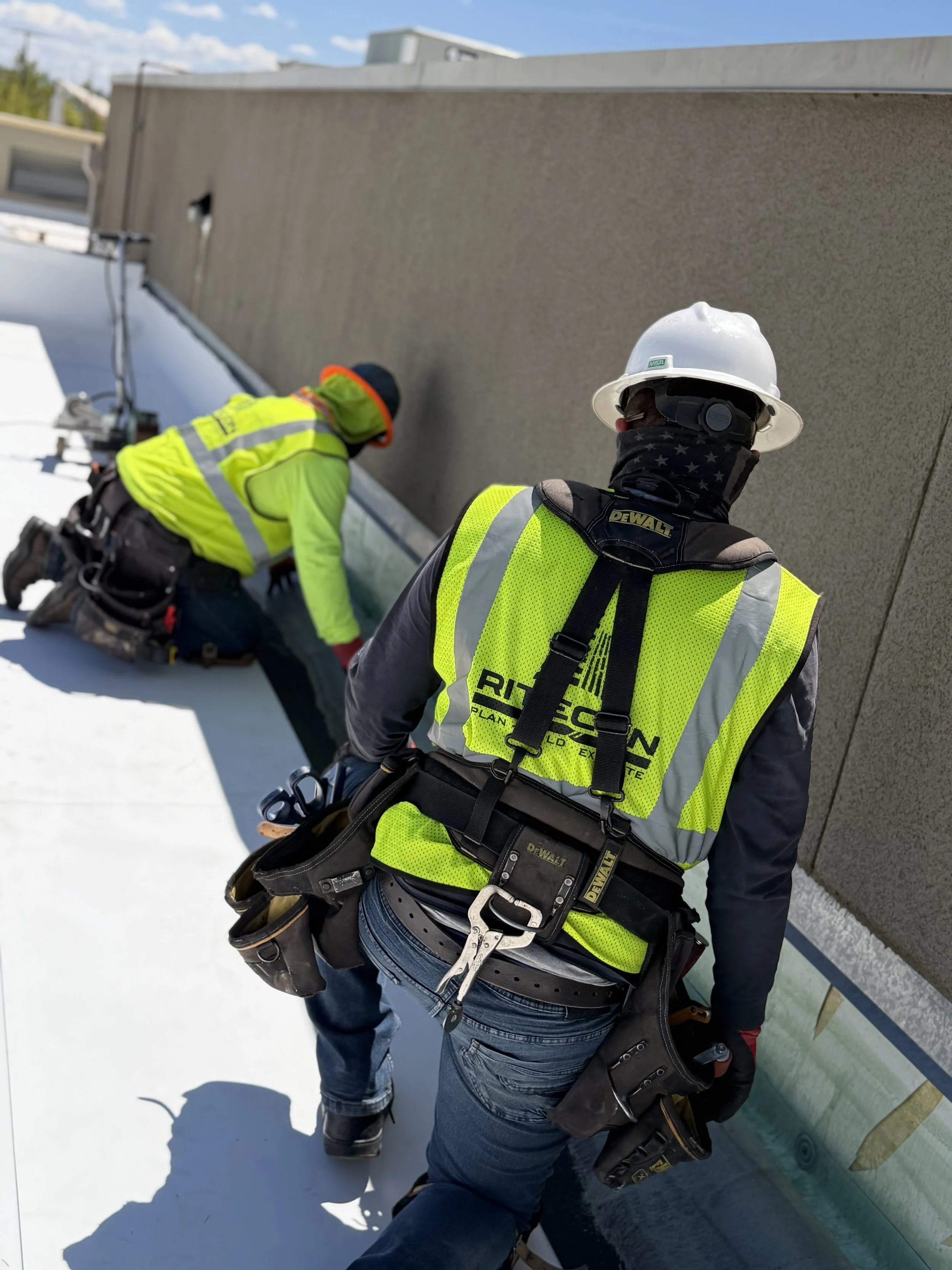 Two construction workers, wearing yellow safety vests and white hard hats, are working on the roof of a building during the daytime. One worker is kneeling, and the other is standing, both focused on their tasks.