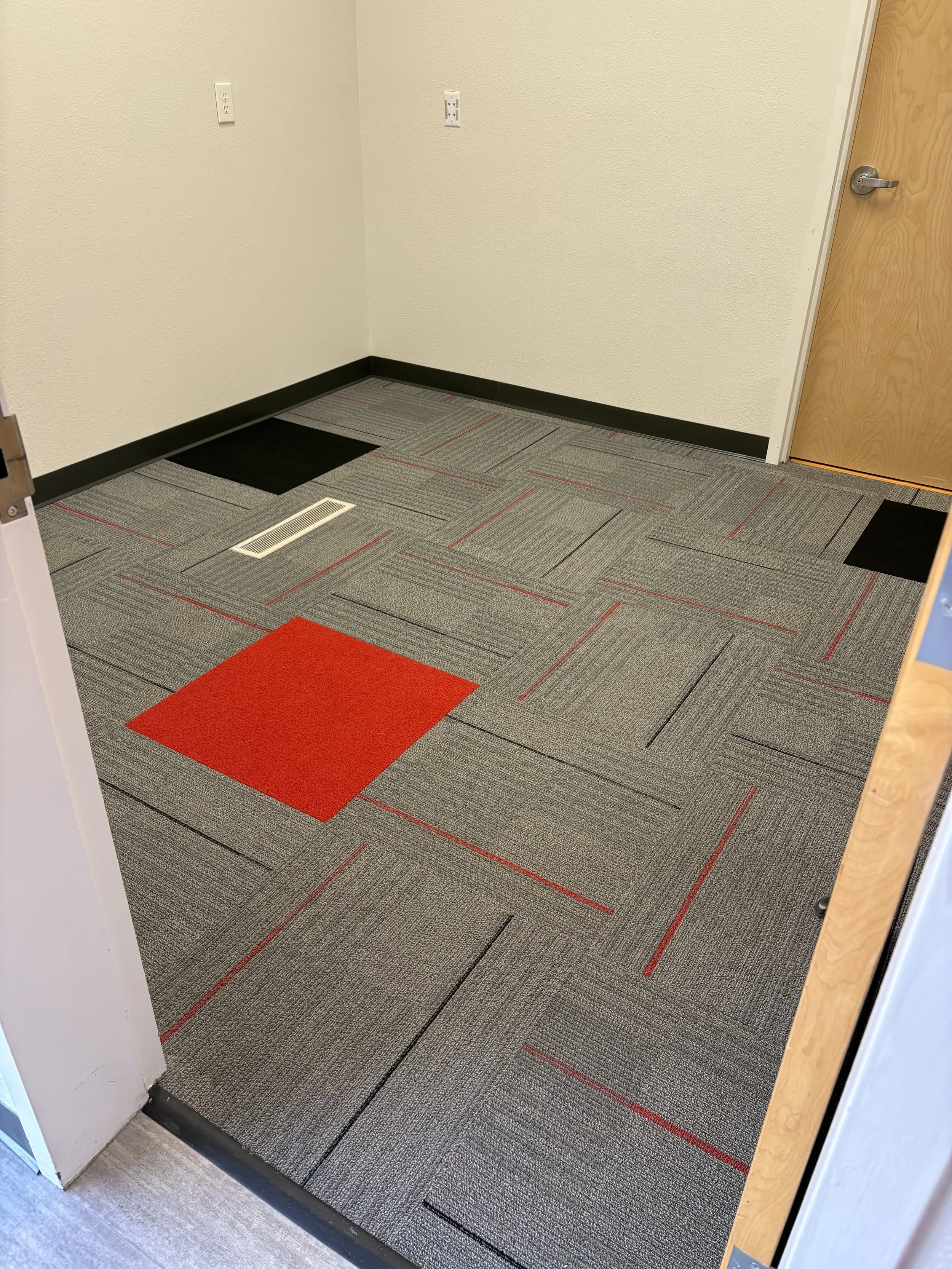 An office or hallway corner with beige textured walls, an electrical outlet, and a wooden door. The floor is covered with gray carpet tiles with red accents and some black tiles, and there is a red carpet tile near the edge.
