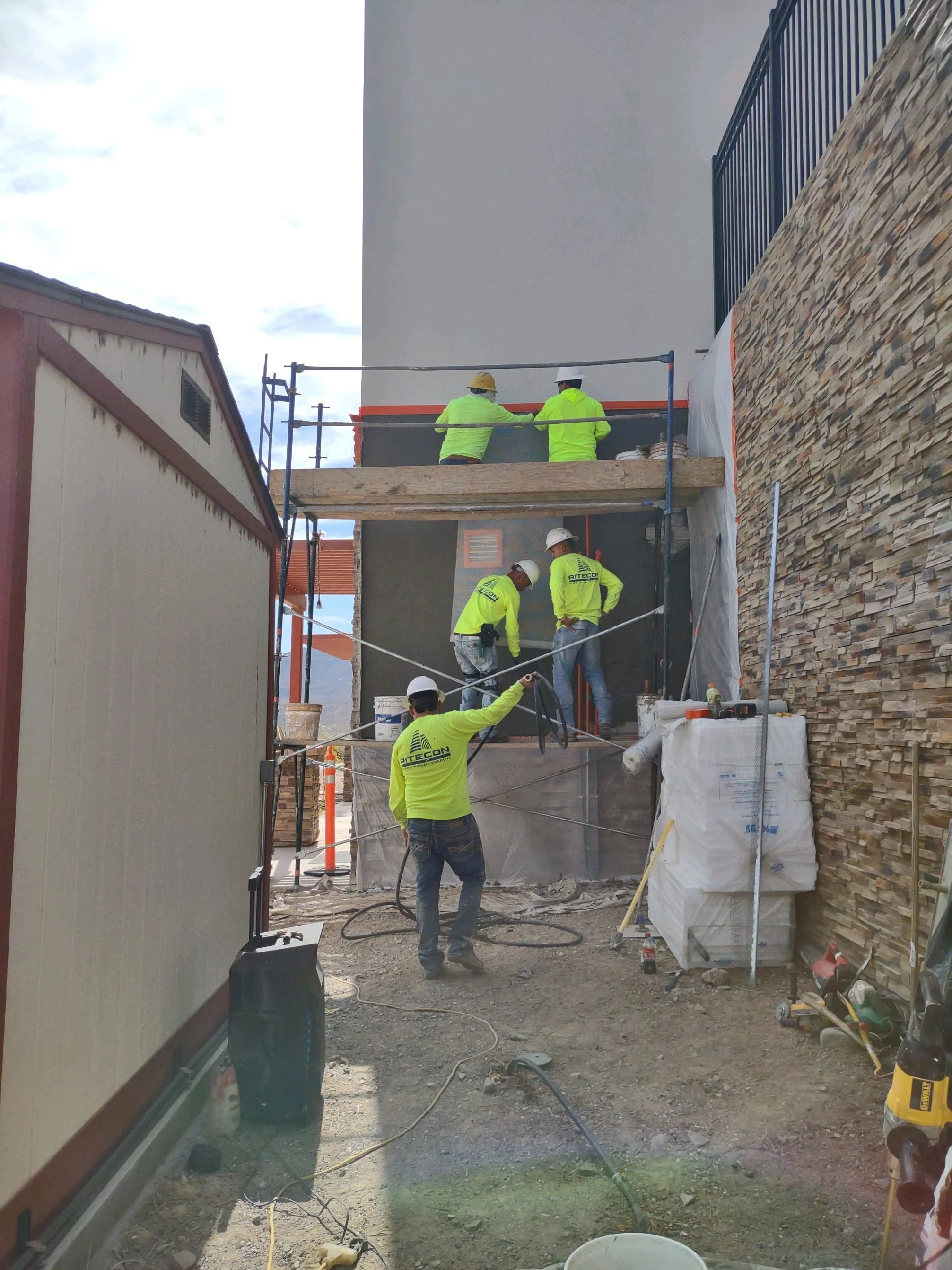 Construction workers wearing safety helmets and high-visibility jackets working on a building exterior, with scaffolding and construction materials around.