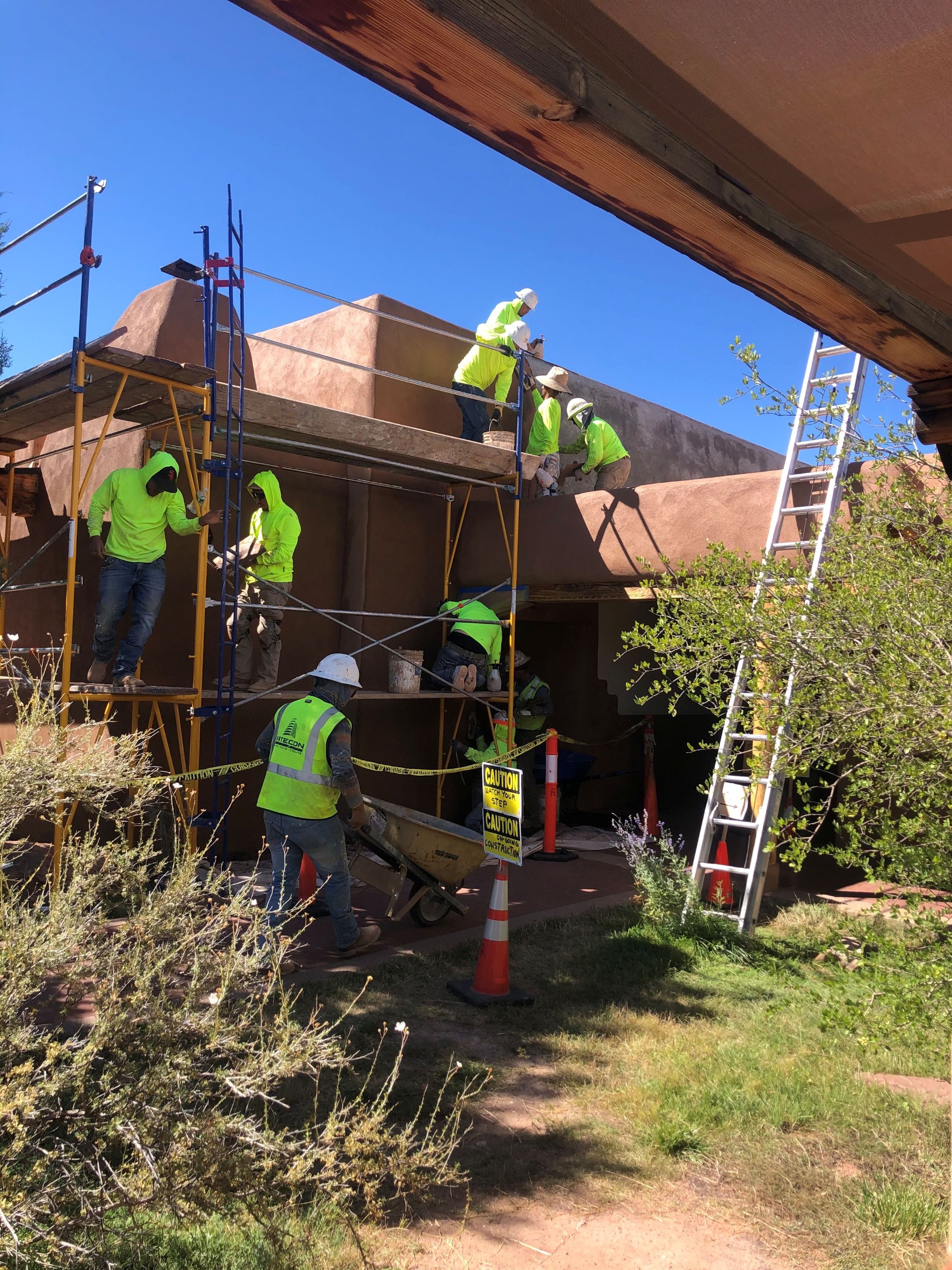 Construction workers in neon yellow safety clothing and white helmets working on building a structure, with scaffolding, ladders, and caution signs, outdoors under a clear blue sky.