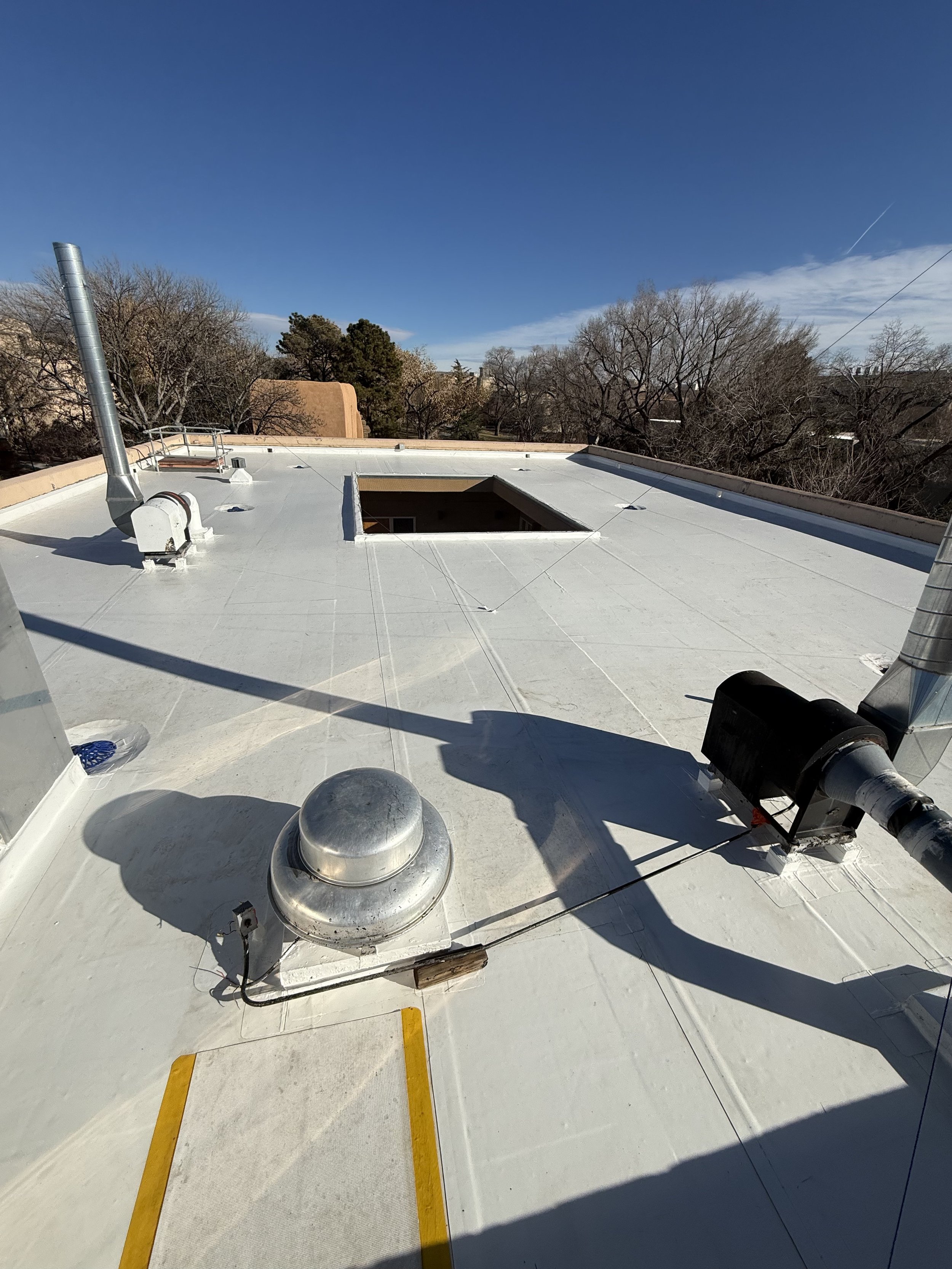 View of a flat commercial rooftop with vents, pipes, an open skylight, and yellow painted lines, under a clear blue sky with some clouds and trees in the background.