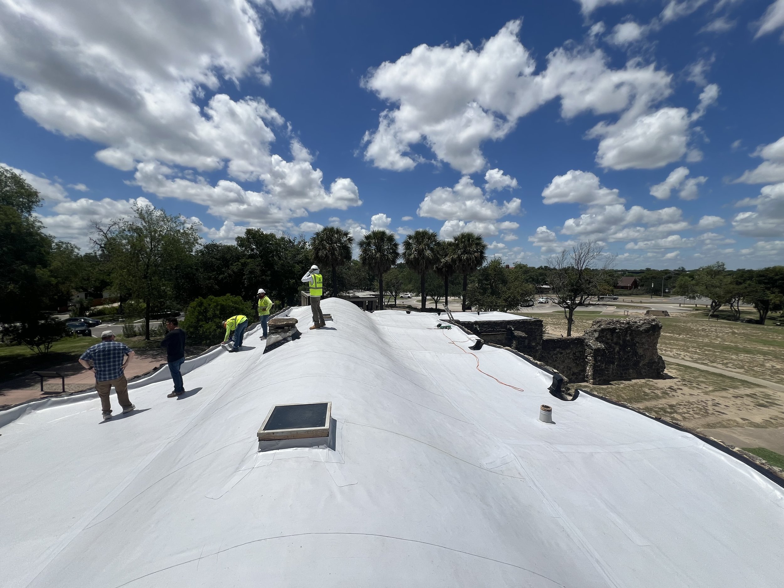 Construction workers in yellow vests and helmets working on a roof covered with a white membrane, with a partly cloudy sky overhead and trees and parking lot in the background.