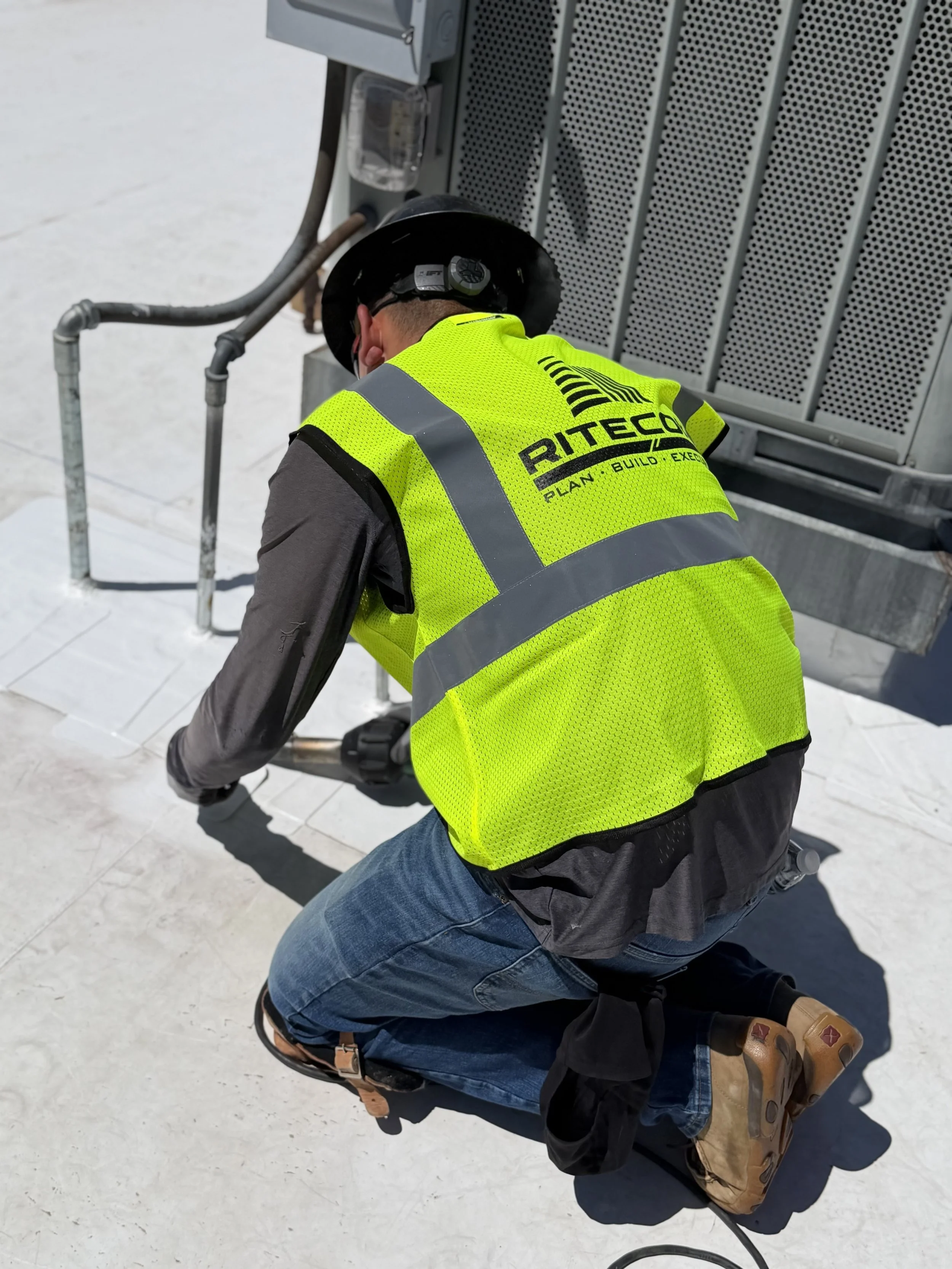 A worker wearing a yellow safety vest and a black hard hat kneeling on a white surface, working on an industrial air conditioning unit or HVAC system.