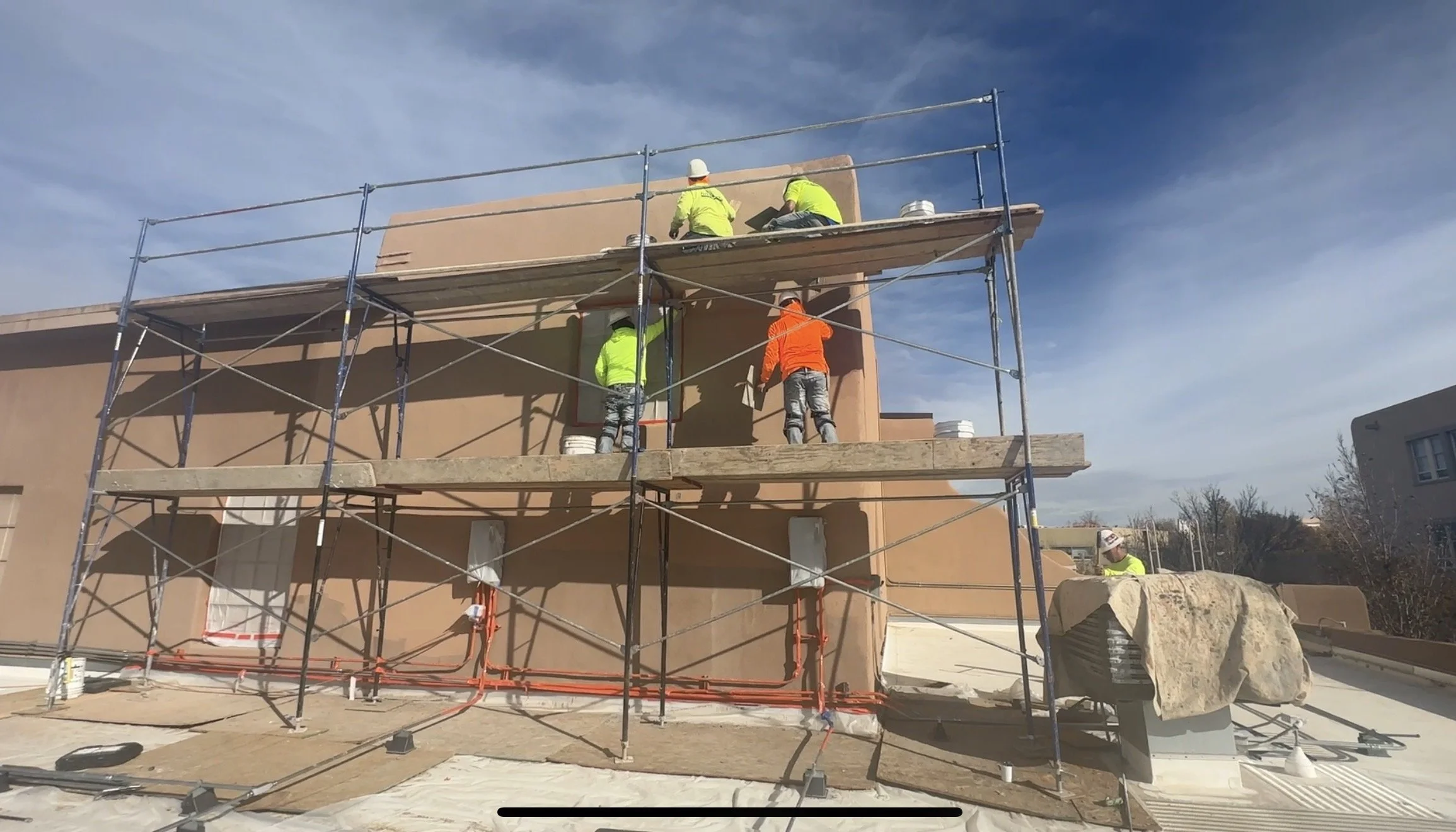 Construction workers on scaffolding working on a building's upper wall, with tools and safety gear, under a blue sky.