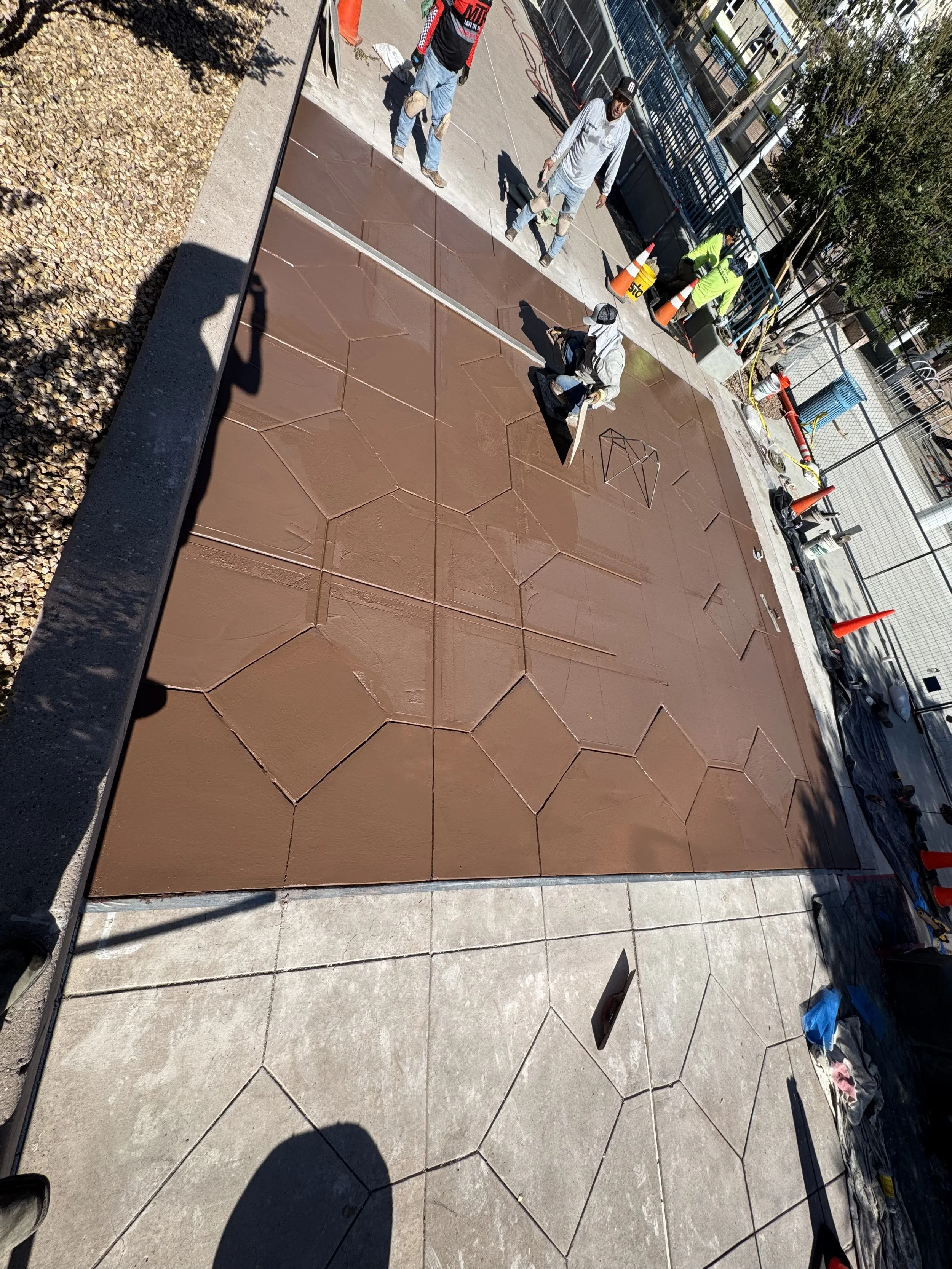 Workers applying brown concrete sidewalk panels on a city street, with construction supplies and safety cones nearby.
