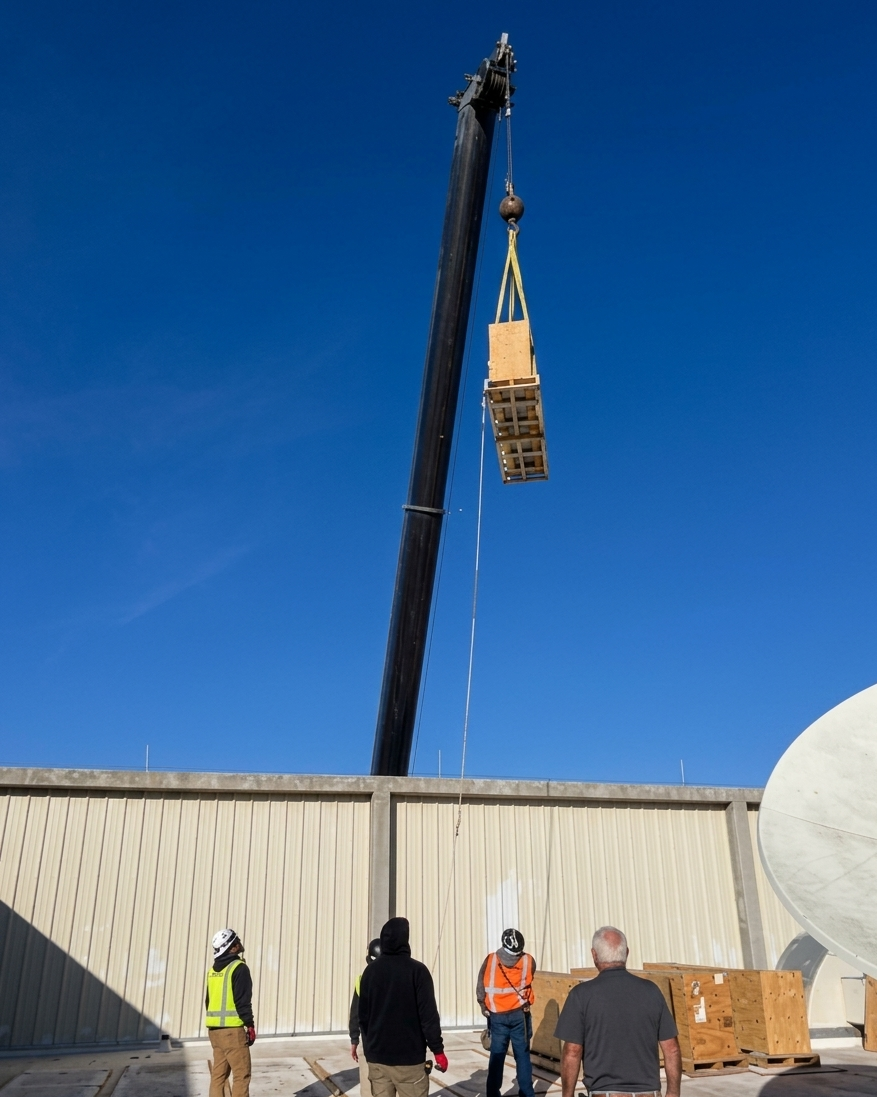 Construction workers observing a crane lifting a wooden pallet onto a building site against a clear blue sky.