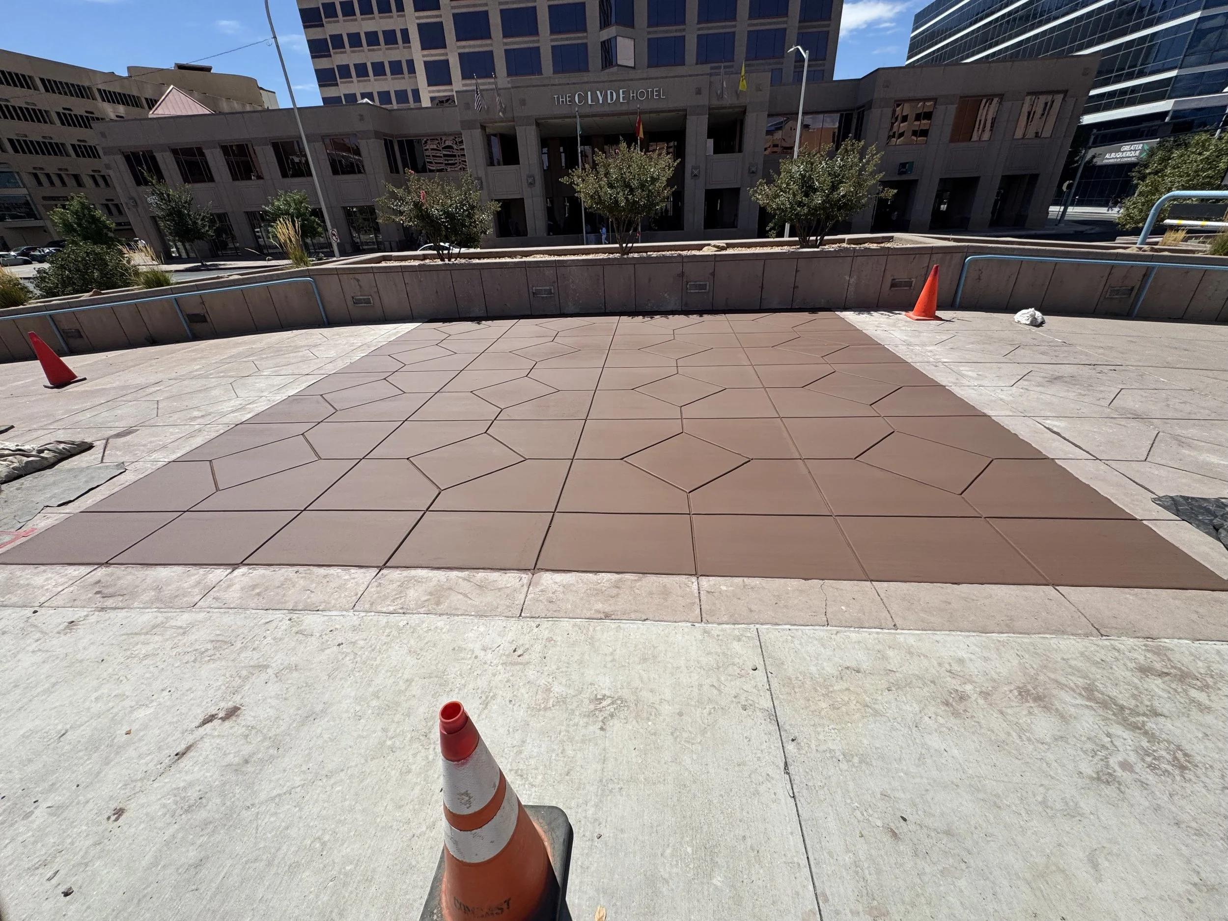 Construction cones on sidewalk in front of a building with trees and glass windows, with taller buildings in the background.
