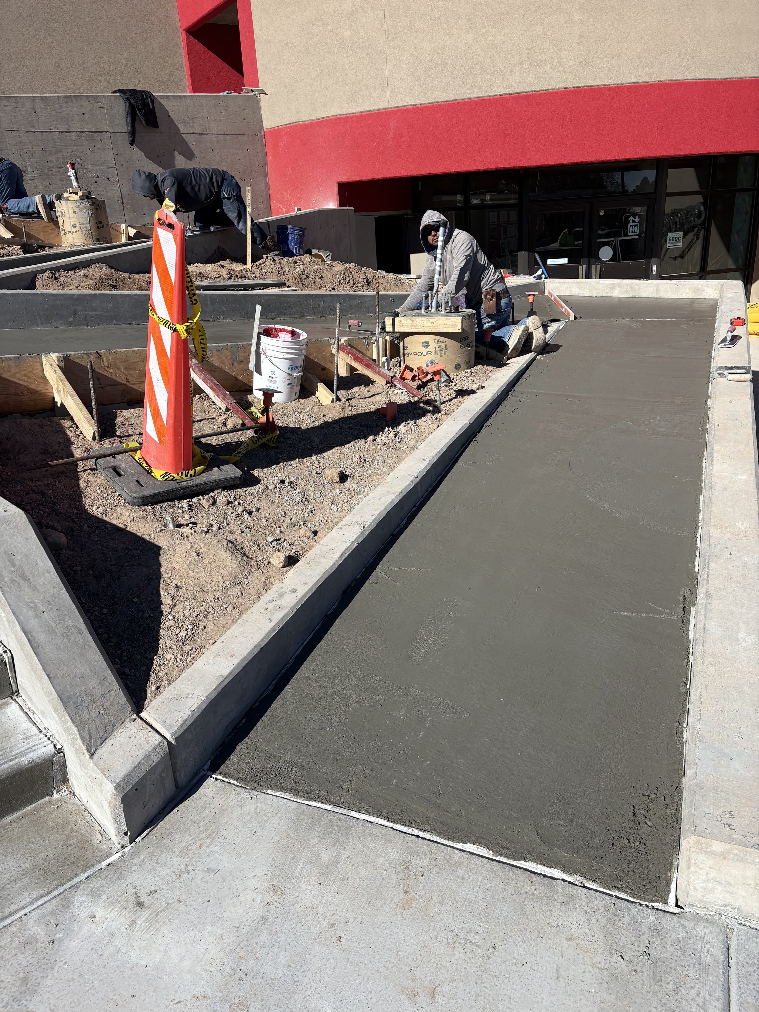 Construction workers working on a concrete sidewalk and ramp outside a building with red and beige walls.