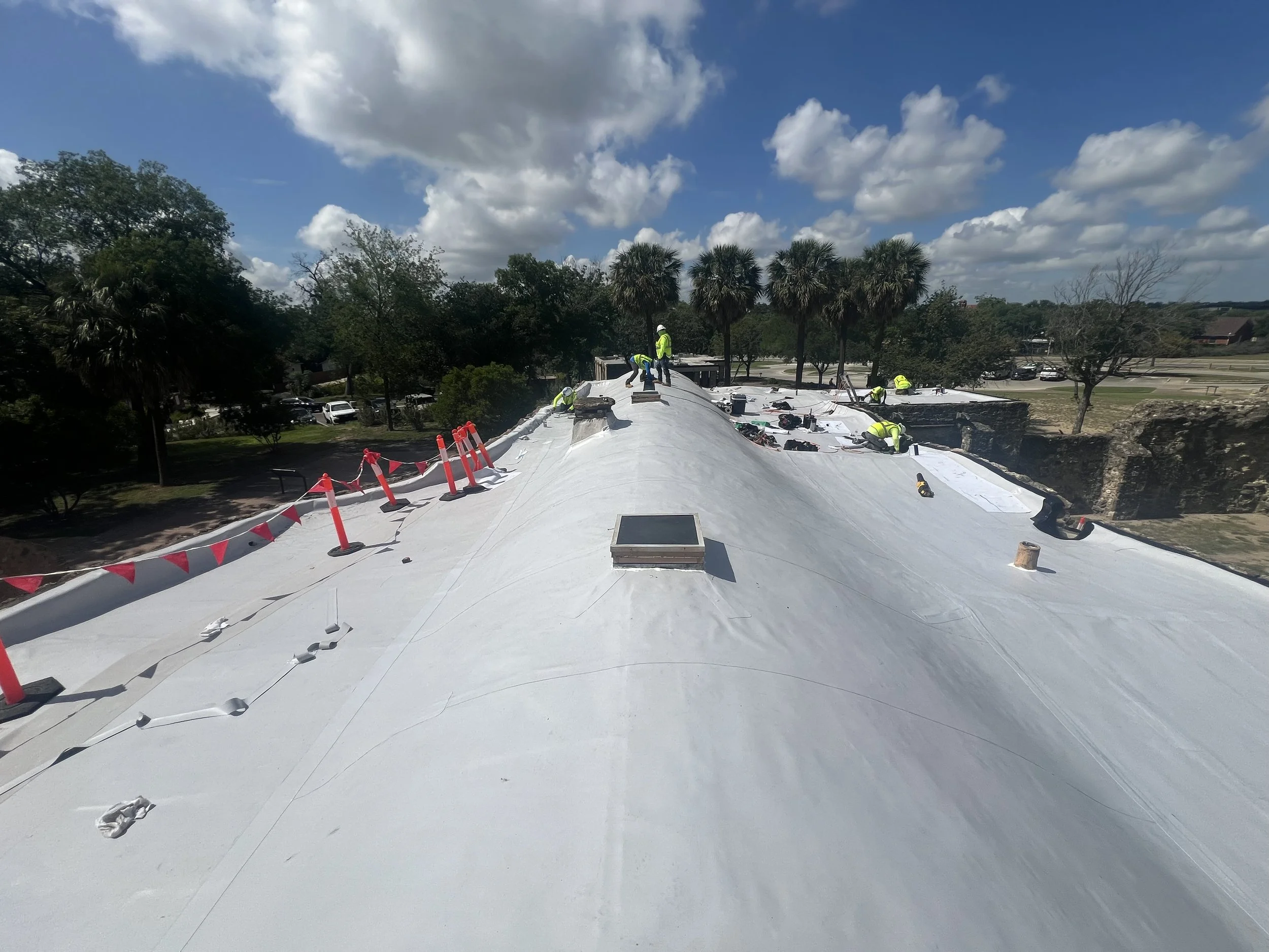 Construction workers on a flat rooftop with white waterproofing material, surrounded by orange safety cones and caution tape, with trees and a parking lot in the background under a partly cloudy sky.