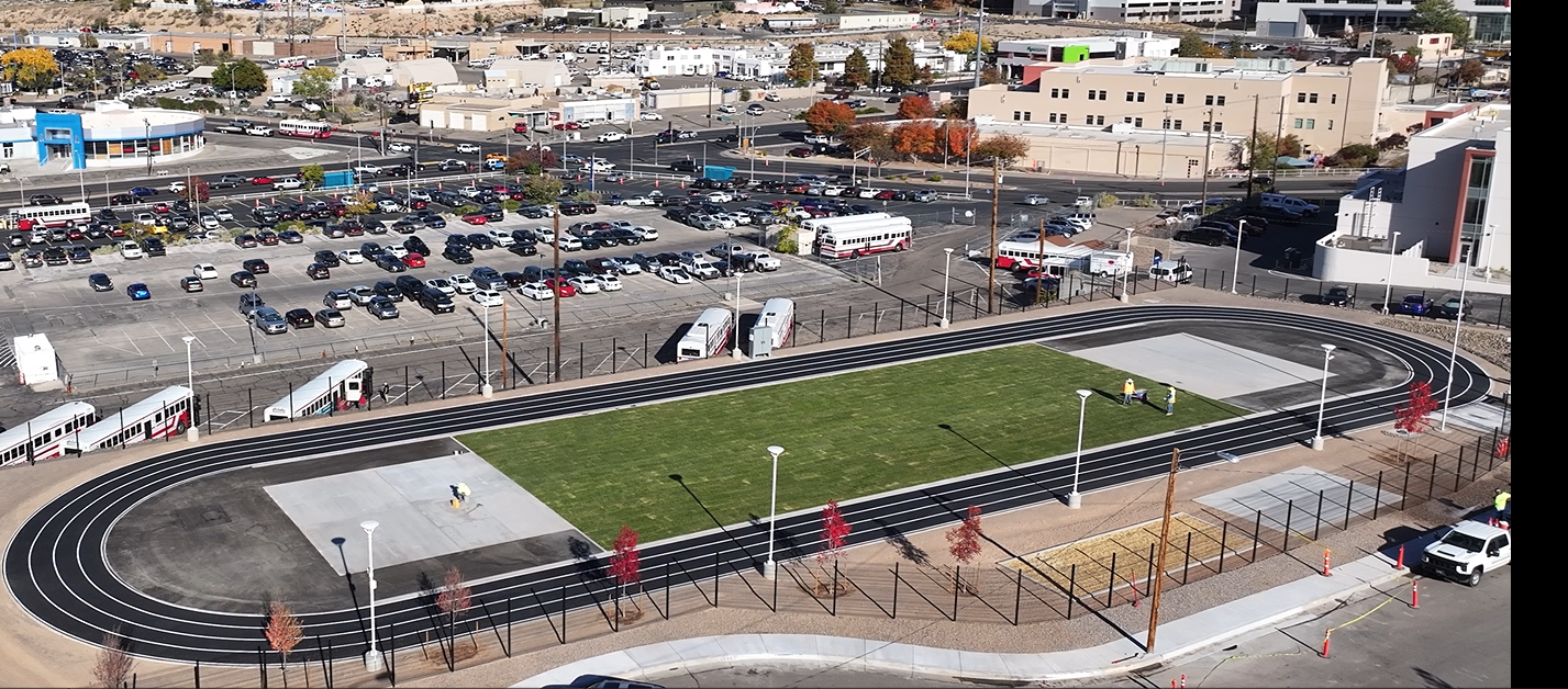 An outdoor track and field area with synthetic running tracks surrounding two sections of grass and concrete pad, situated in a parking lot with parked cars and surrounding buildings.