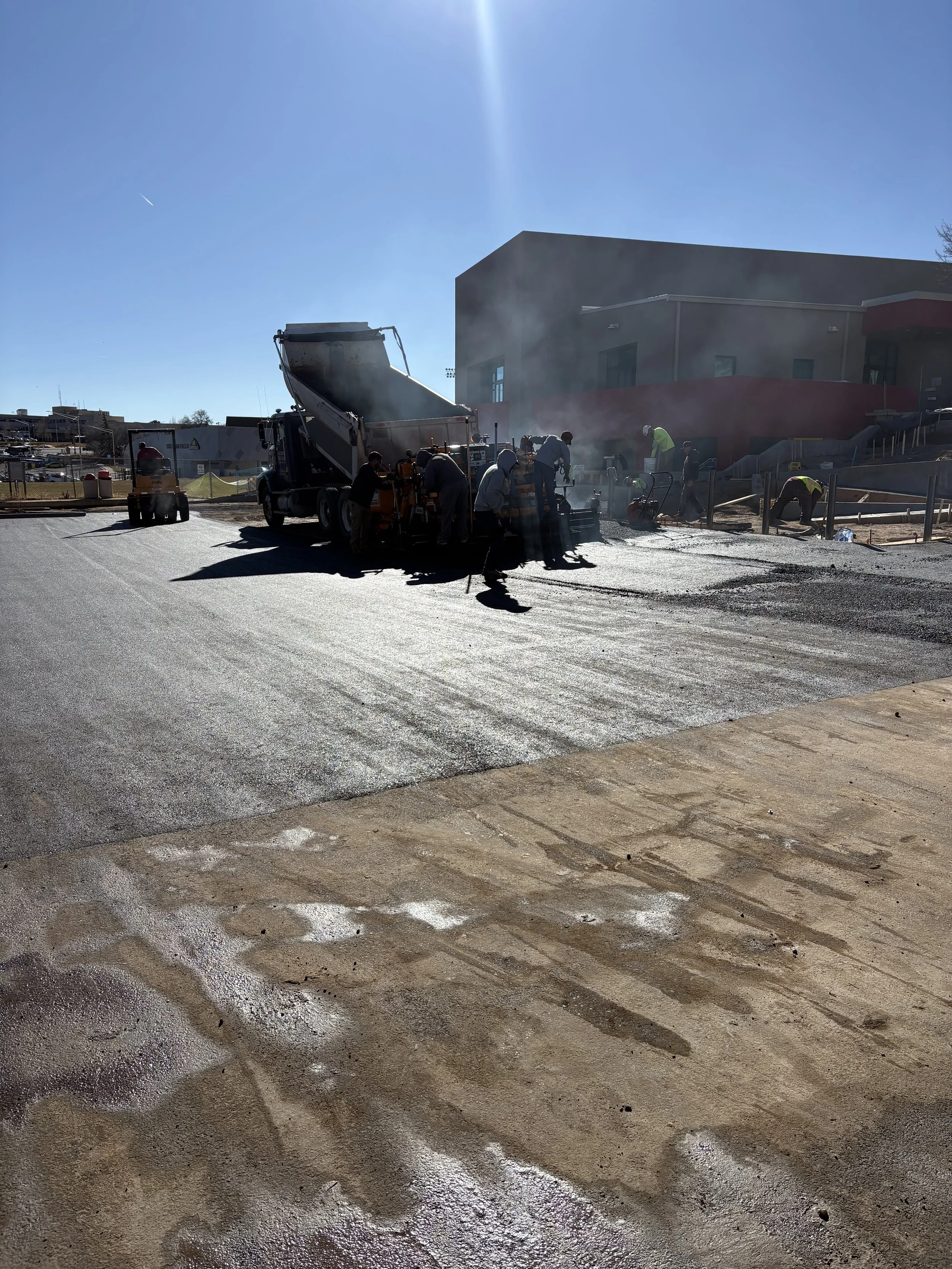 Construction workers paving a road with concrete, with a building in the background under a clear blue sky.