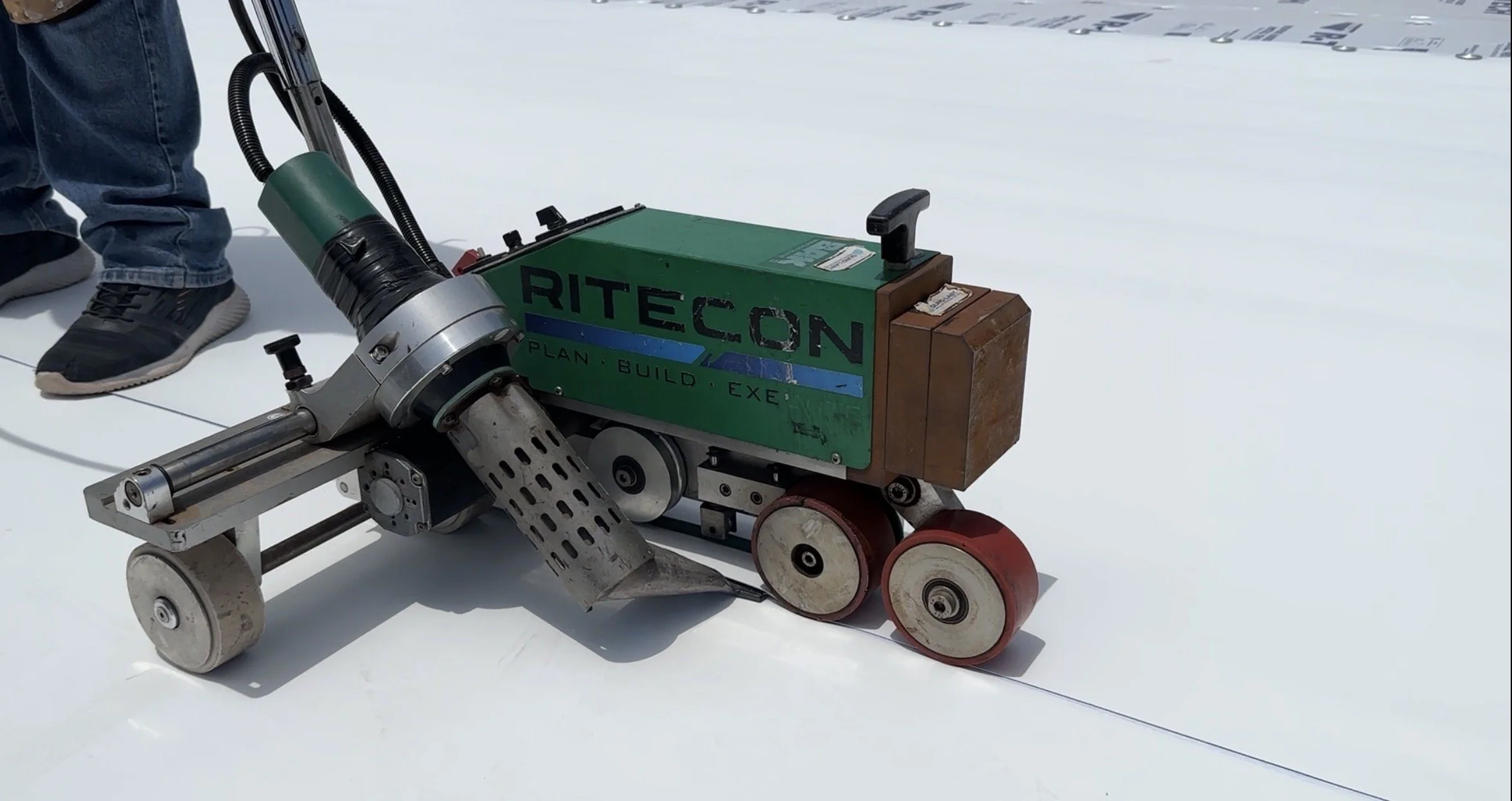 A robotic device with wheels on a white floor, connected to a green box labeled 'RITECON,' with the words 'PLAN, BUILD, EXECUTE' on it. Part of a person's leg and shoe are visible in the background.