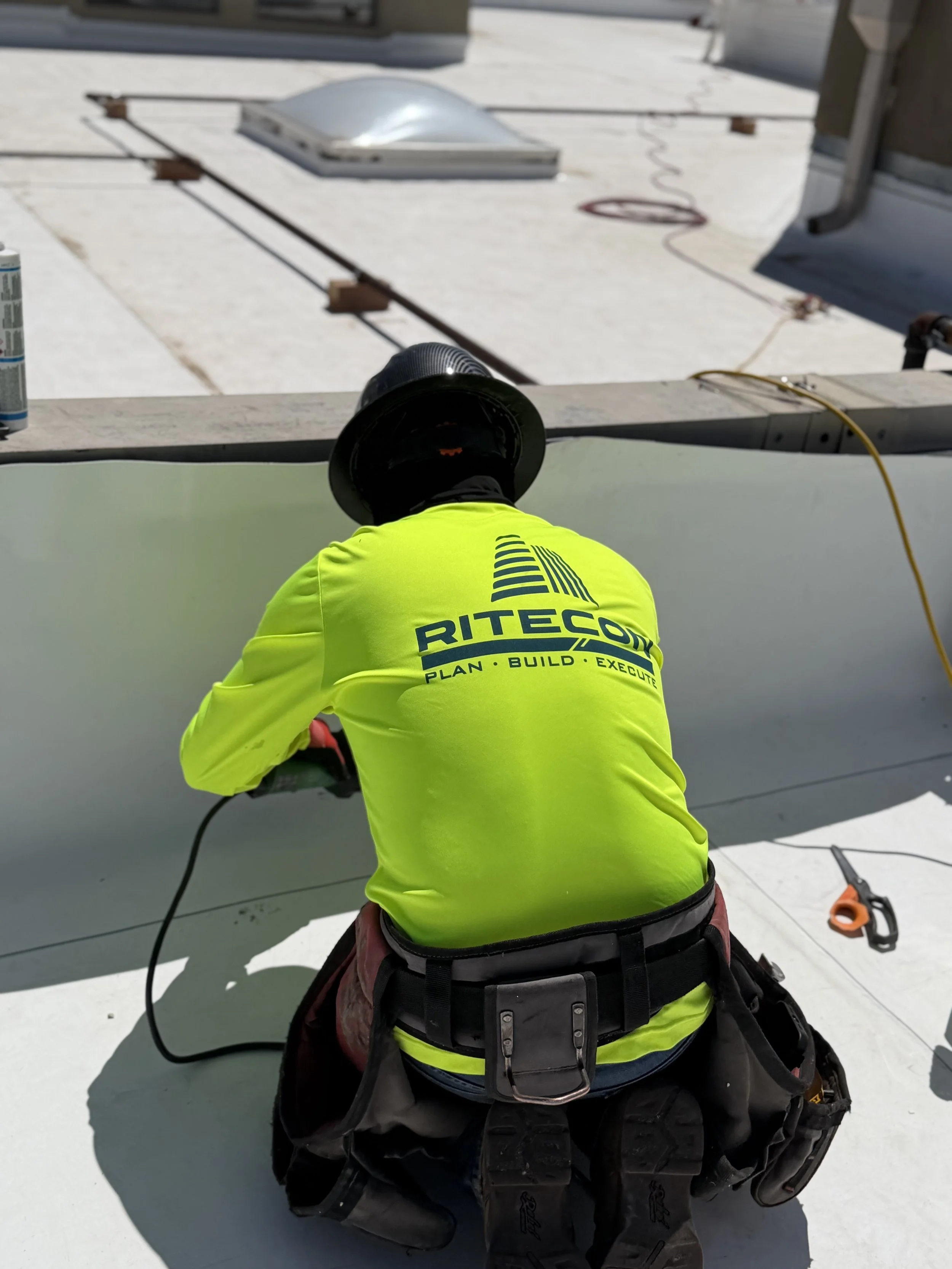 A worker wearing a bright yellow safety jacket, a black helmet, and a tool belt is kneeling on a flat roof. The worker has the word 'RITECON' on the back of the jacket. The roof has a white surface with a skylight and some construction tools and wiri