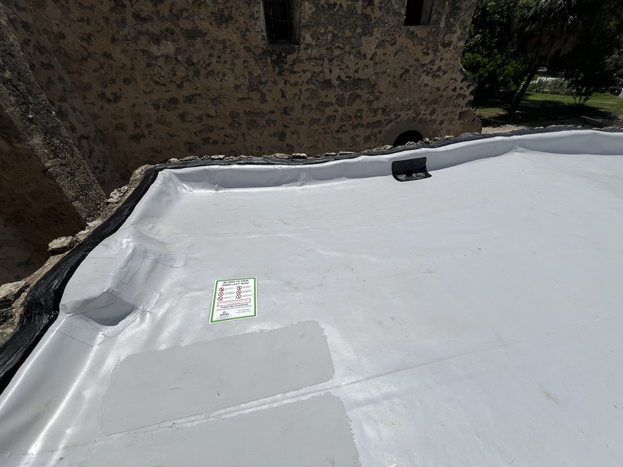 Photo of a white roofing membrane on a flat roof with a black border and a green and white warning label in the center. In the background, there is an old stone wall, trees, and a grassy yard.