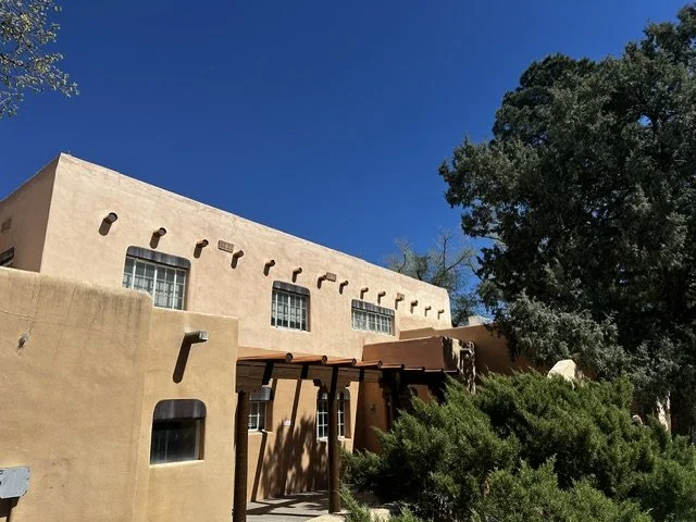 A southwest-style building with adobe walls, small windows, and a wooden shaded entrance, surrounded by green bushes and a large tree under a clear blue sky.