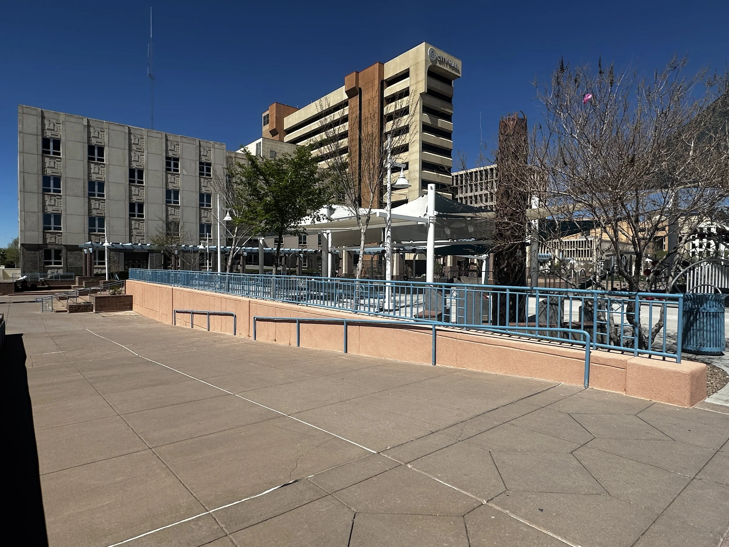 Empty plaza with a paved walkway, leafless trees, blue metal benches, and a covered seating area, with multistory buildings in the background under a clear blue sky.