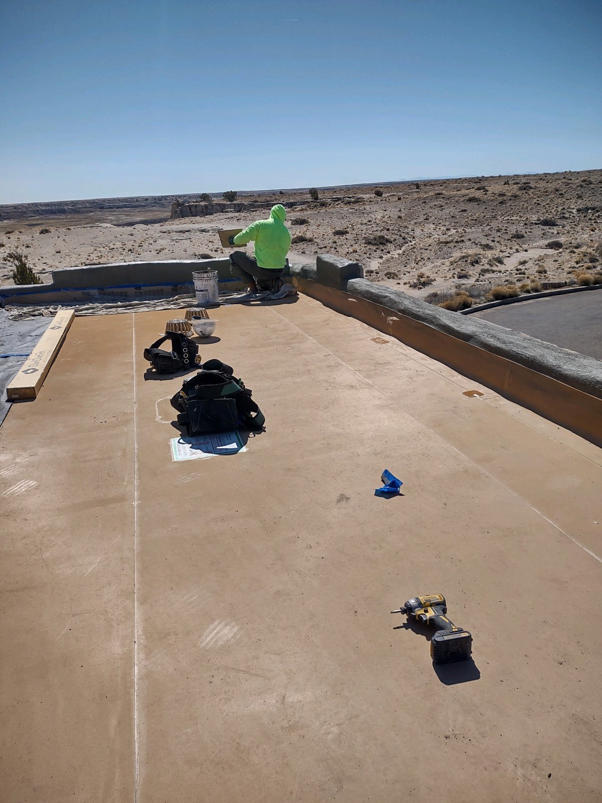 A construction worker in a bright green hoodie working on a rooftop with desert landscape in the background. Tools, bags, and construction supplies are nearby on the flat surface.