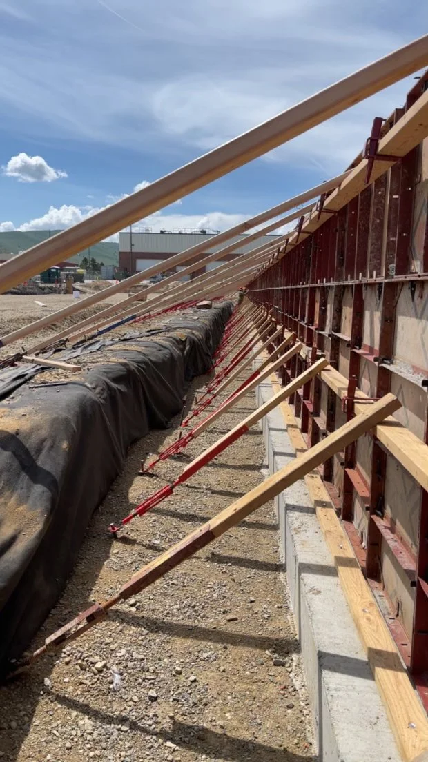 Construction site with wooden formwork and braces for concrete wall, gravel ground, and partly cloudy sky.