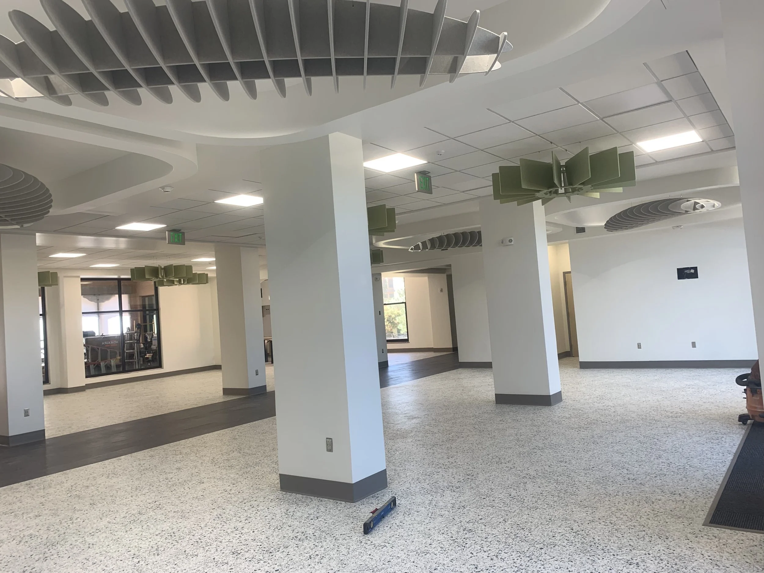 A modern, spacious interior with white walls and ceiling, suspended ceiling tiles, and rectangular light fixtures. There are decorative green panels hanging from the ceiling and multiple large vent diffusers. Several white columns support the ceiling