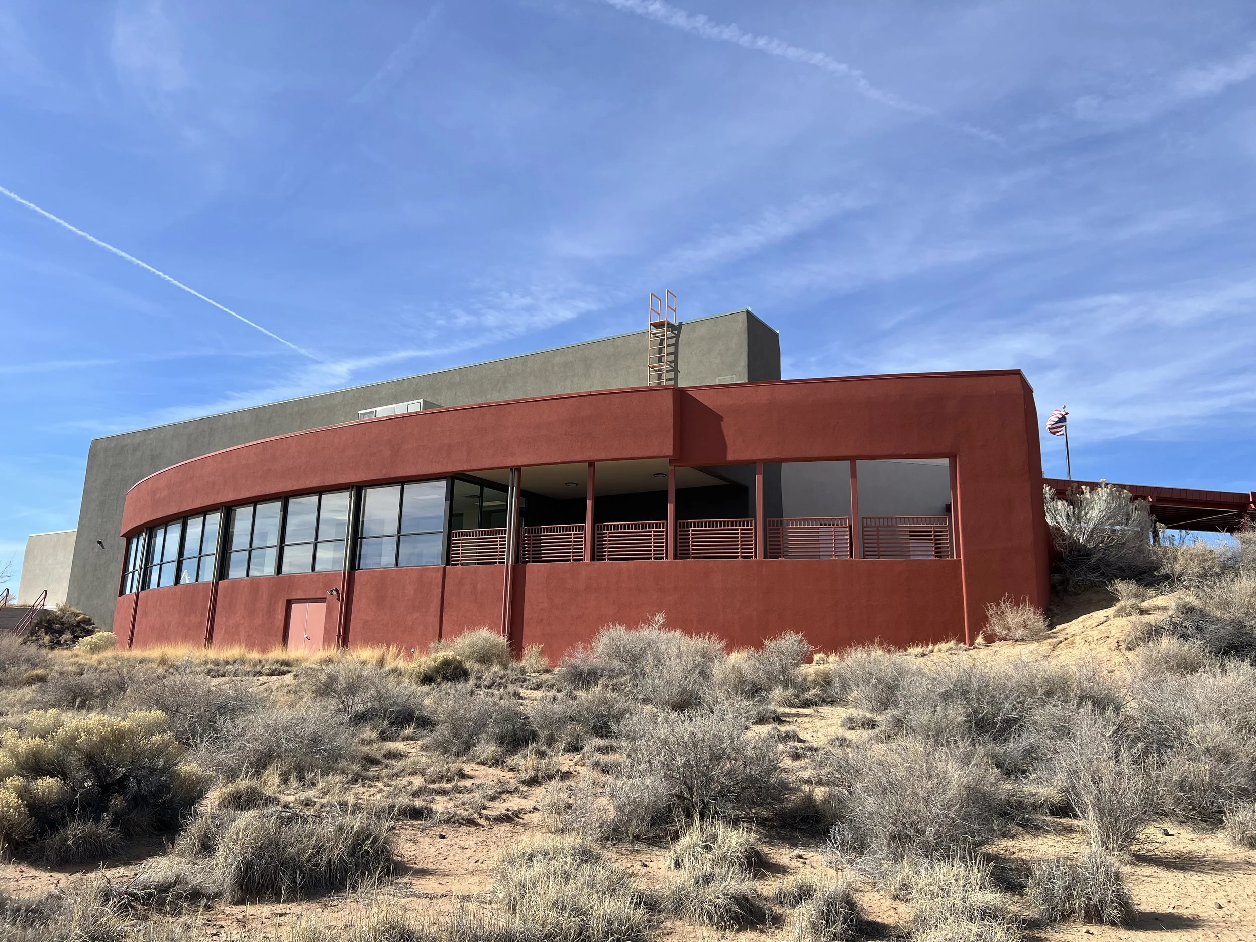 A modern building with a curved red exterior and large glass windows, situated in a dry, shrub-covered landscape under a clear blue sky with some wispy clouds.