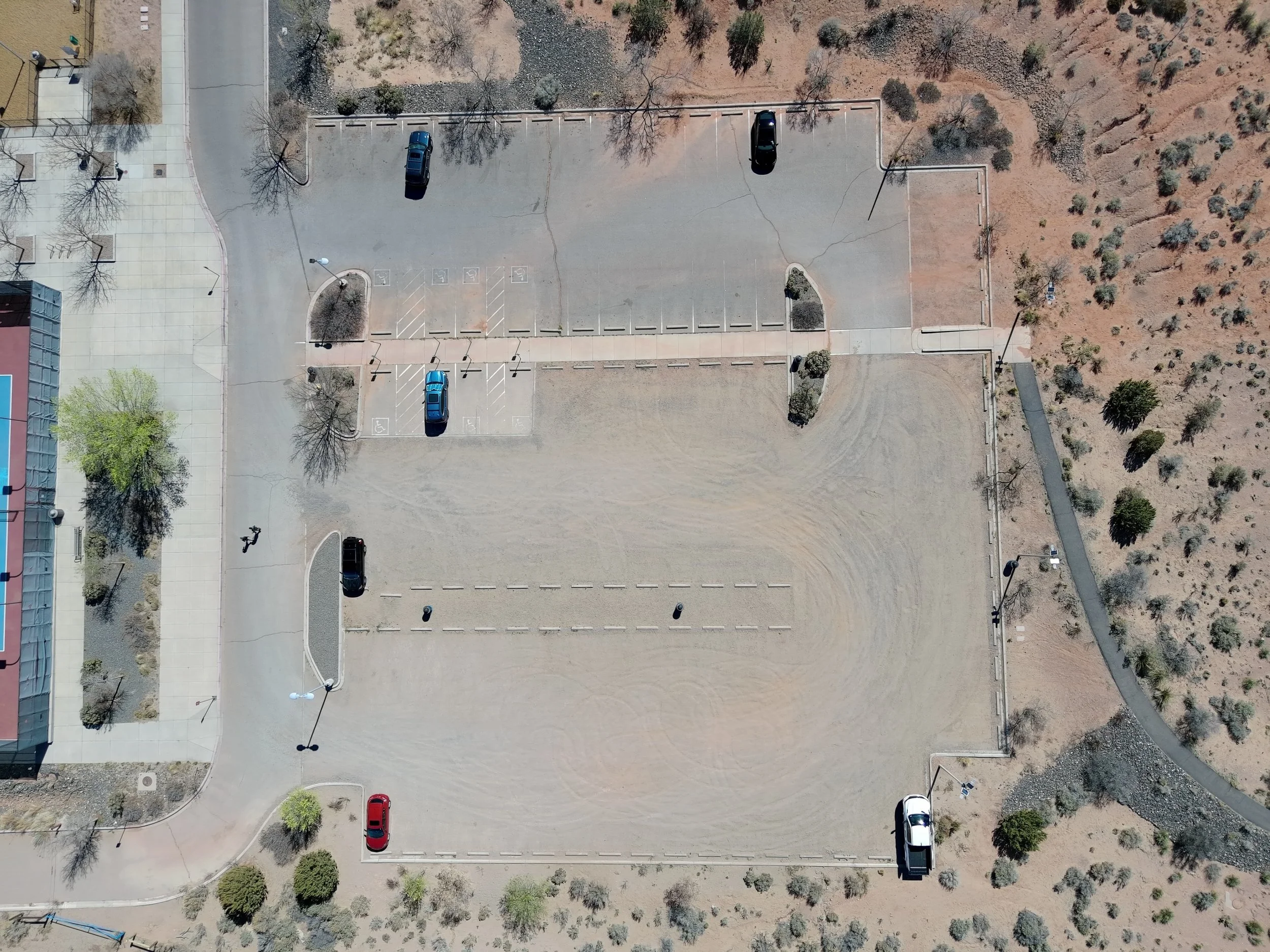 An aerial view of a mostly empty parking lot with a few cars parked, surrounded by trees and a sidewalk, with a building to the left and a dry, grassy area to the right.