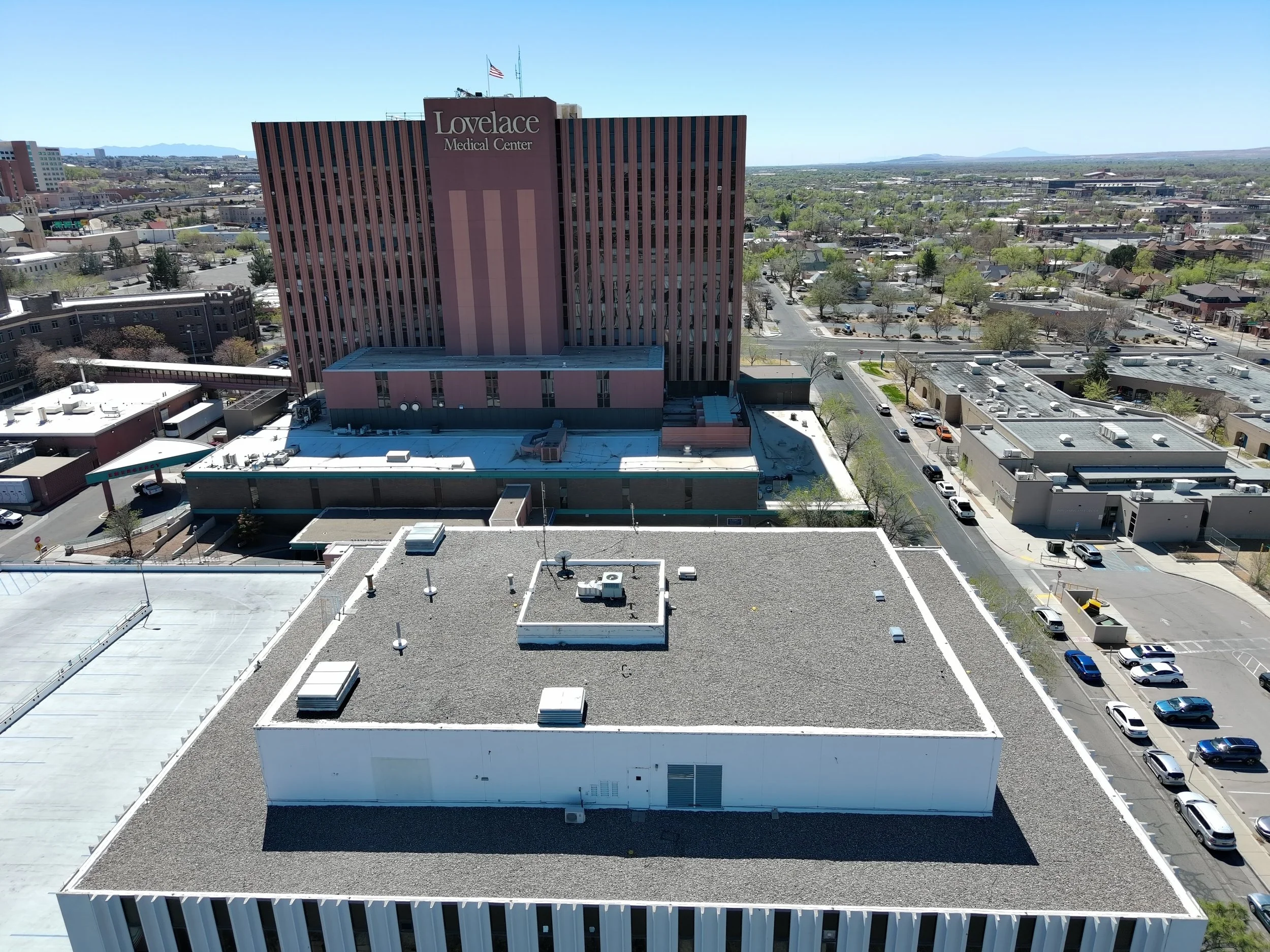 High-angle view of Lovelace Medical Center building with red and beige exterior, surrounded by parking lots, trees, and neighboring buildings in a cityscape, under clear blue sky.