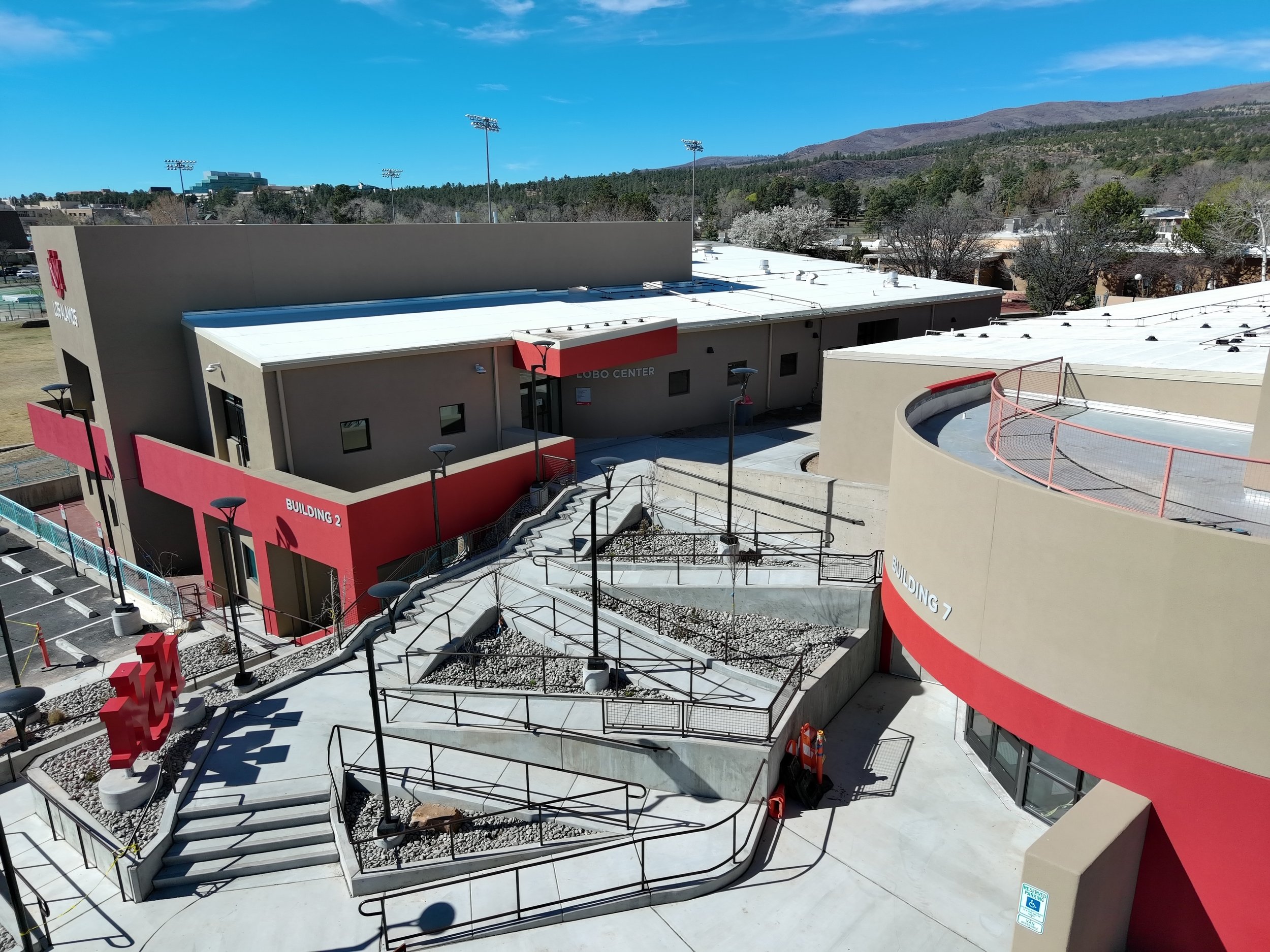 Exterior view of a modern school building with a flat roof, outdoor staircases, and a basketball court, with mountains and trees in the background under a blue sky.