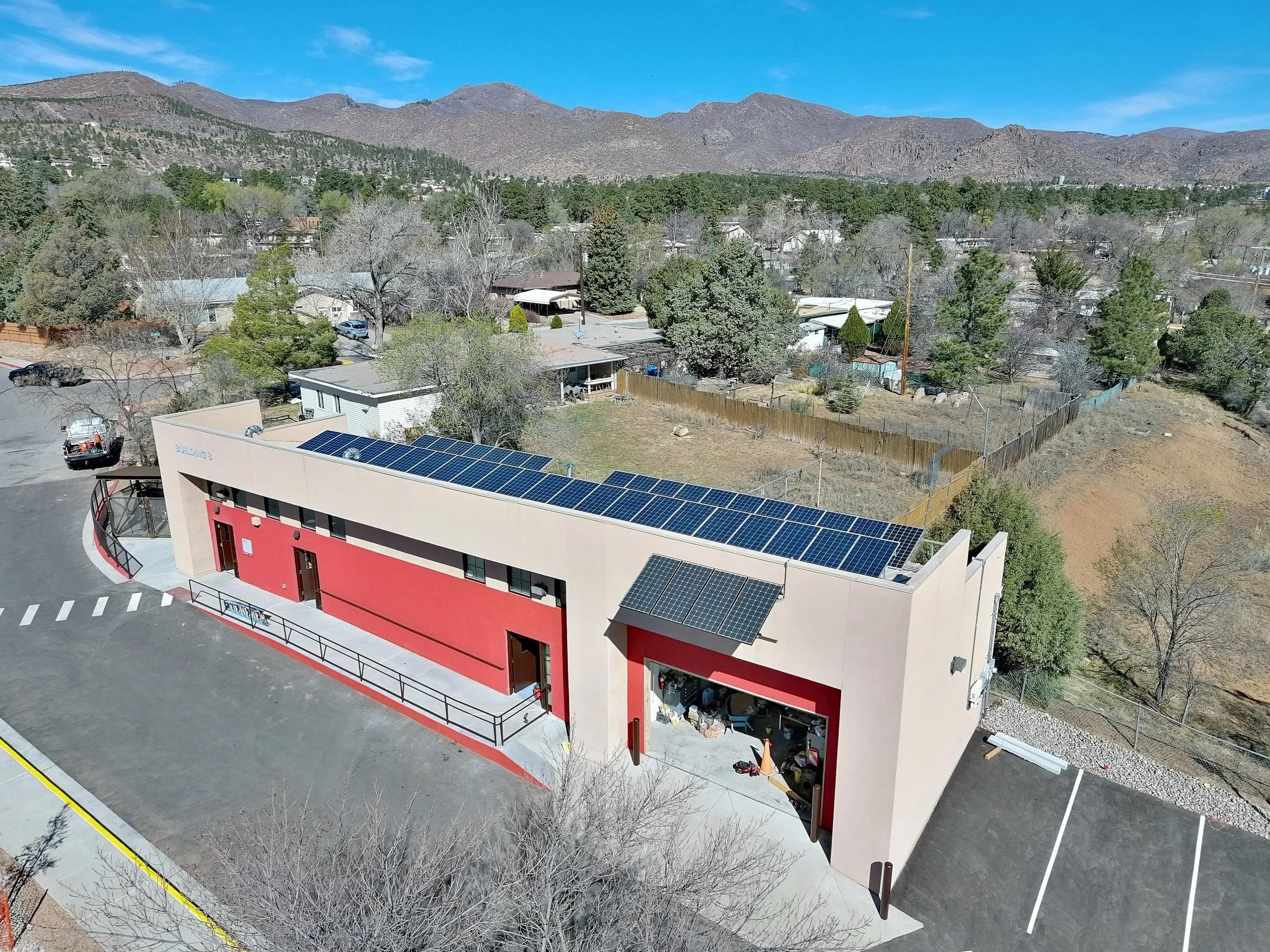 Aerial view of a building with solar panels on the roof, parking lot with cars, trees, and residential area with mountains in the background.
