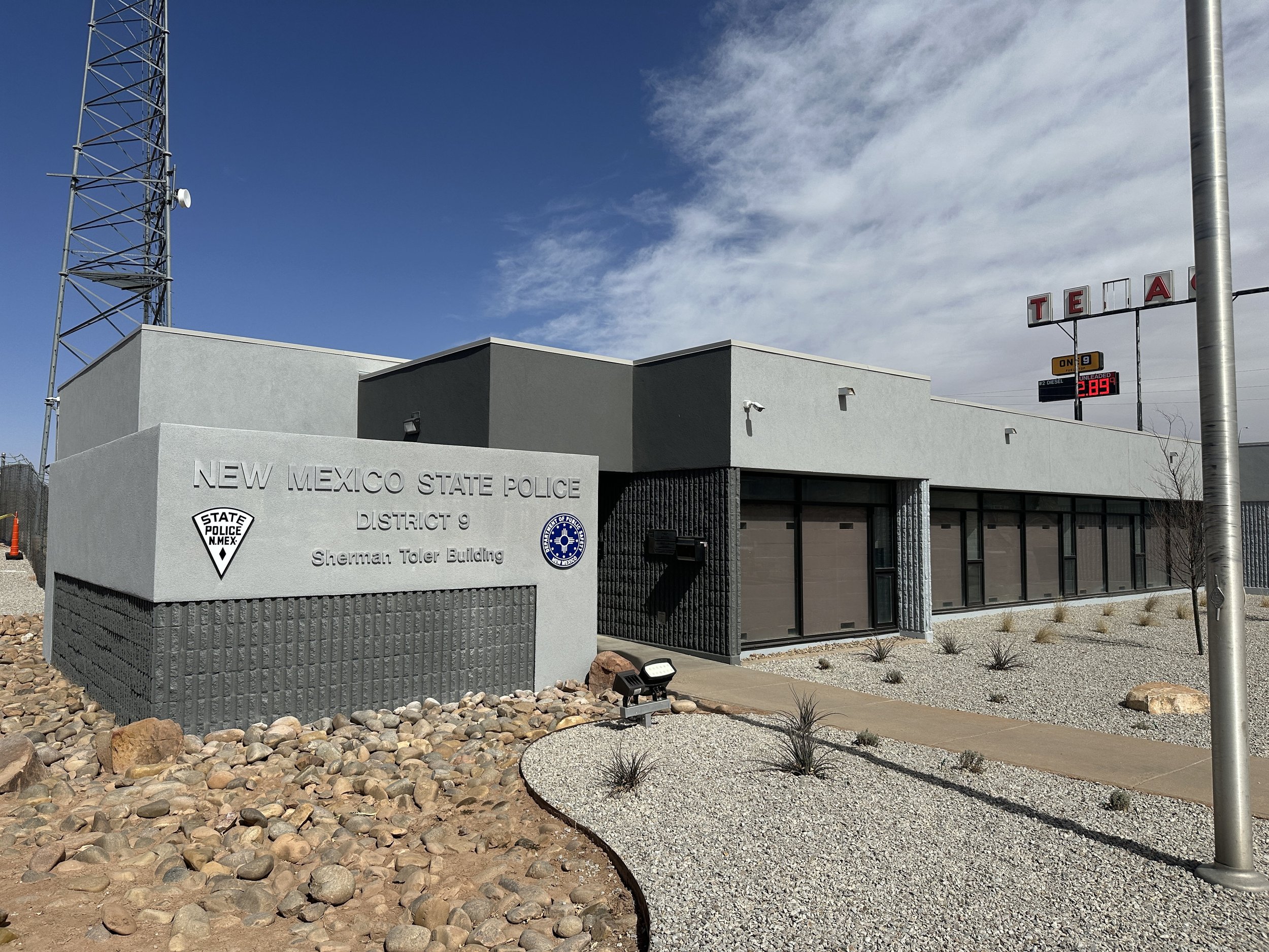 The entrance of the New Mexico State Police Sherman Toler Building, with a sign displaying fuel prices and an outdoor decorative landscaped area with rocks and small plants, under a partly cloudy sky.