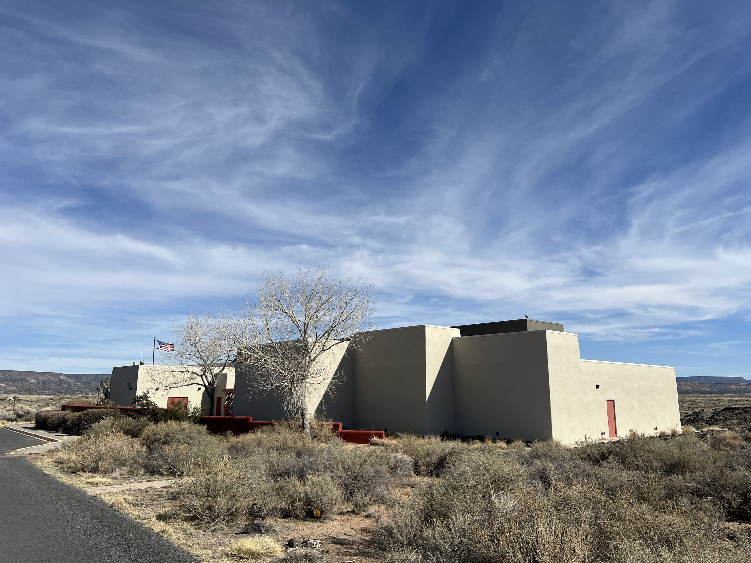 A modern building with light-colored walls, a red door, and a sloped roof, set in a desert landscape with dry bushes and a tree, under a blue sky with wispy clouds.