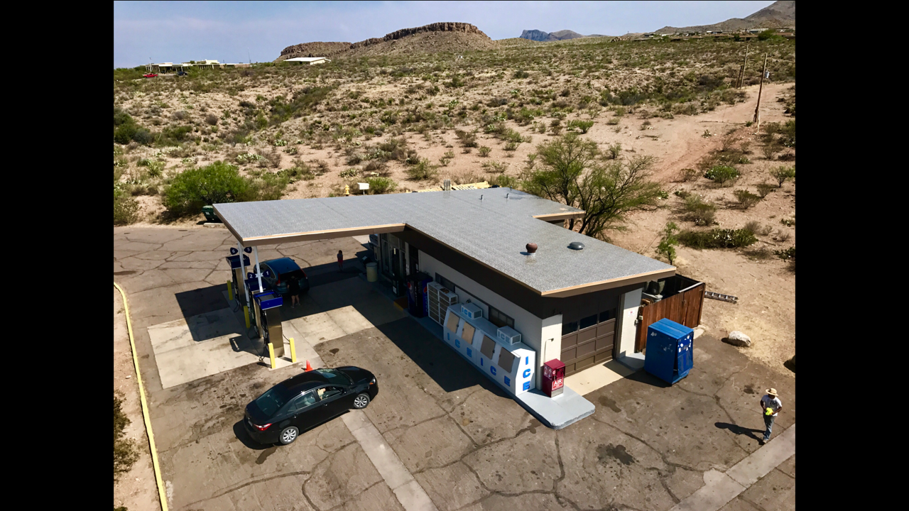 A small roadside convenience store with a car wash in the desert, surrounded by sparse vegetation and distant hills, with two cars parked in front and people nearby.