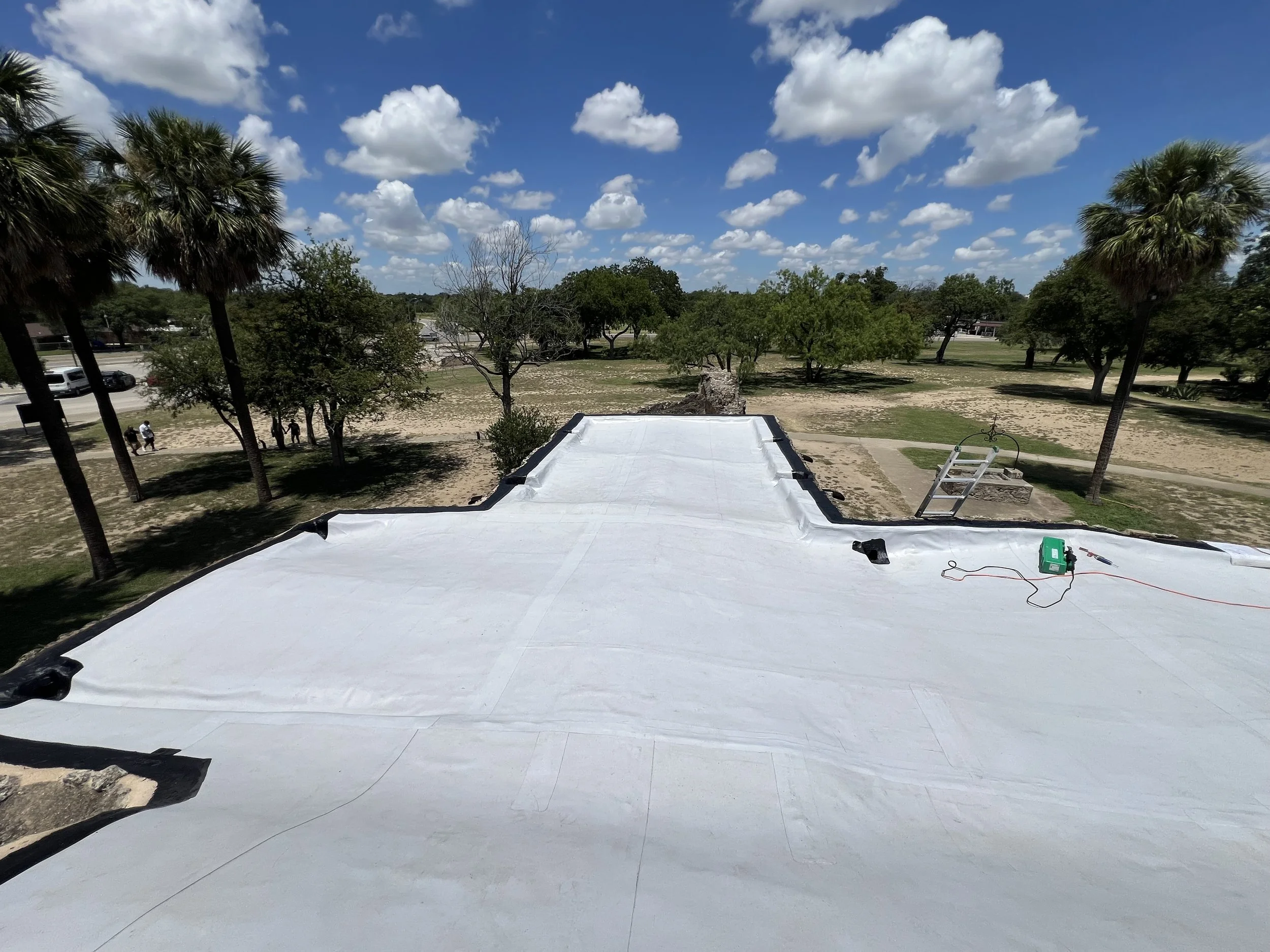 The image shows a rooftop with a white waterproofing layer being applied, surrounded by a landscape with trees, some pedestrians, and a parking lot under a partly cloudy sky.