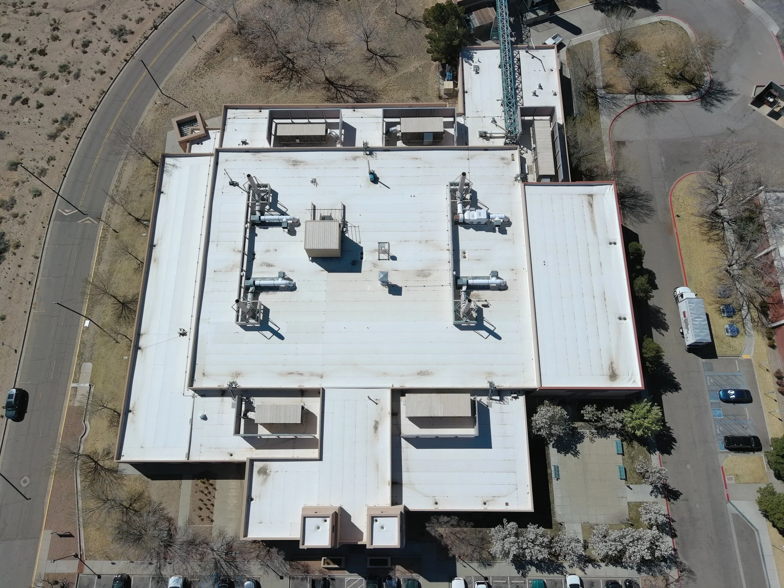 An aerial view of a large white building with multiple vents and air conditioning units on the roof, surrounded by parking lots, trees, and a curved road.