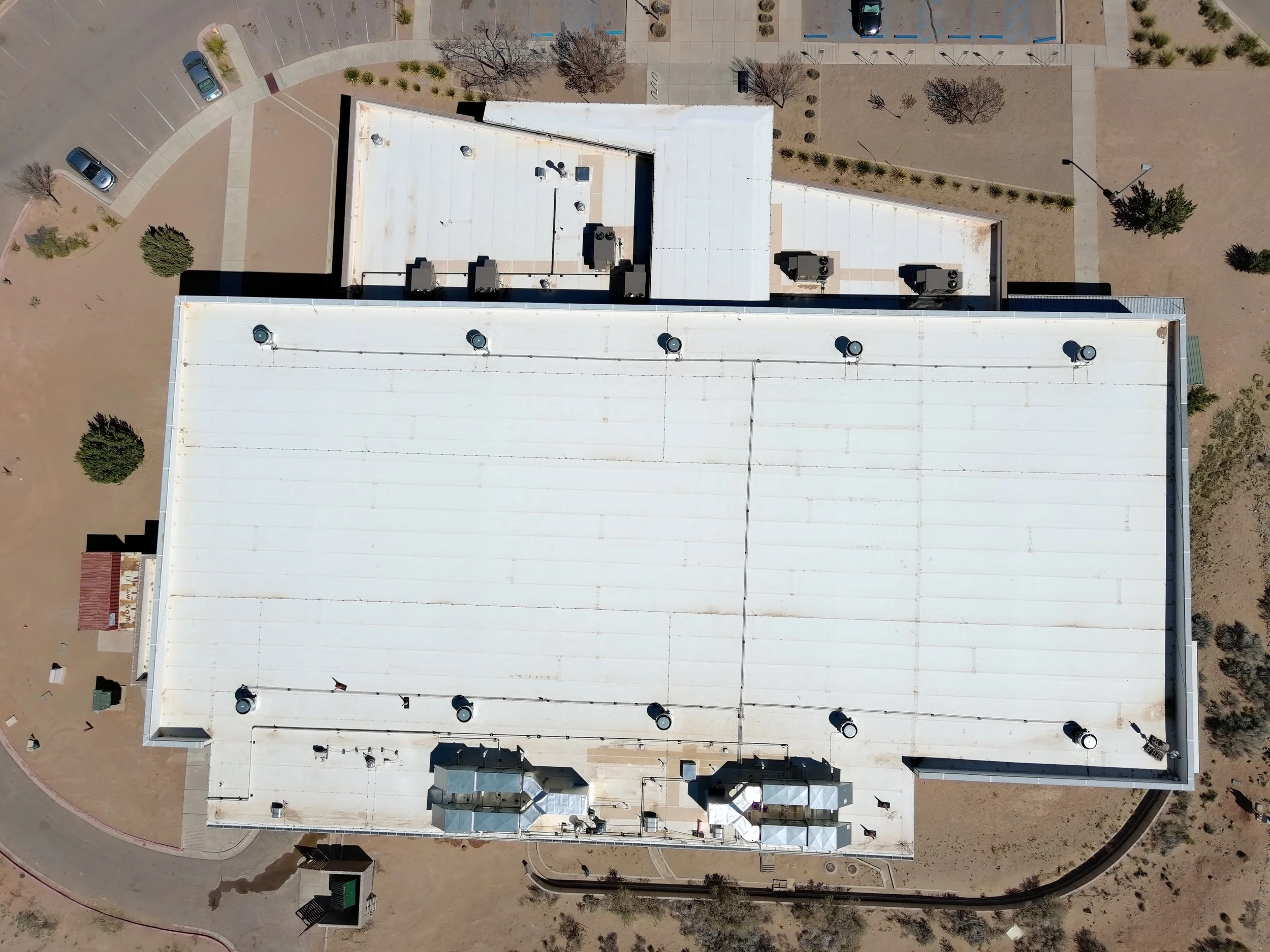 An aerial view of a building with a flat white roof, air conditioning units, and vents. Surrounding the building are parking lots with a few parked cars, walkways, trees, and landscaped areas. The scene appears to be in a desert or arid environment.