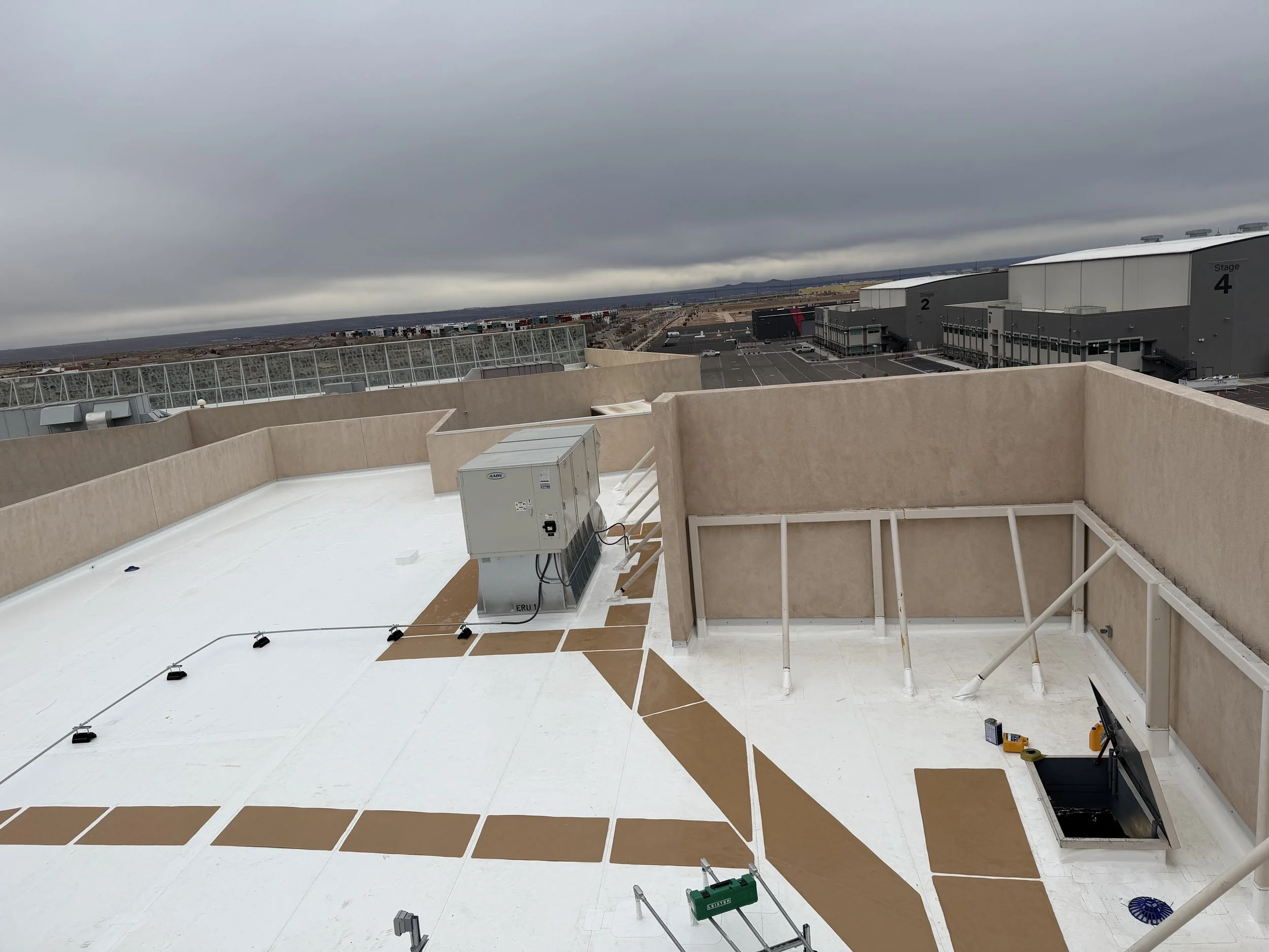 A rooftop with a white surface, beige walls, and various equipment, overlooking an overcast landscape with industrial buildings, roads, and a parking lot. Gray storm clouds cover the sky.