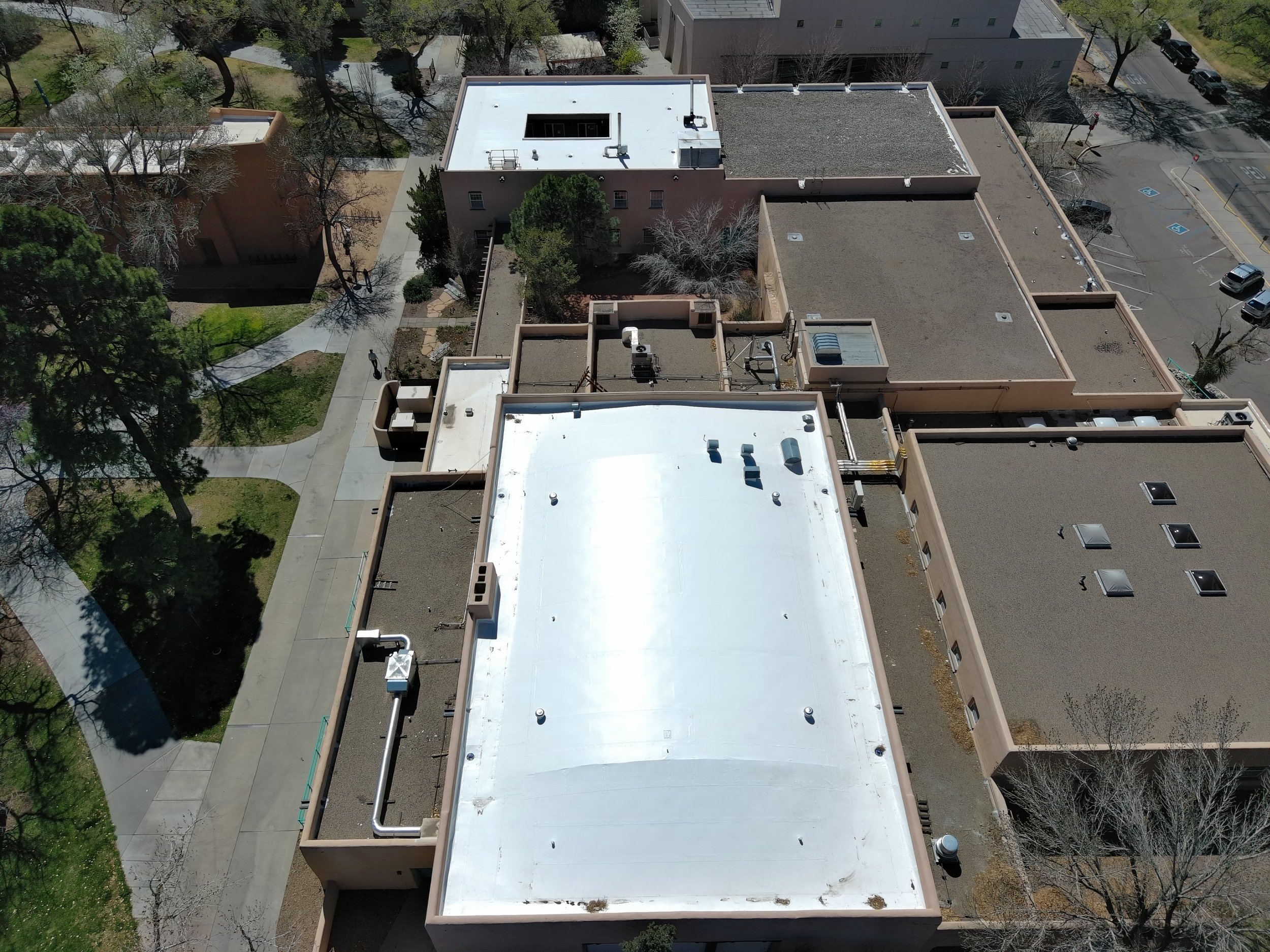 An aerial view of flat rooftops on a building, with a parking lot and trees nearby, in a sunny urban setting.