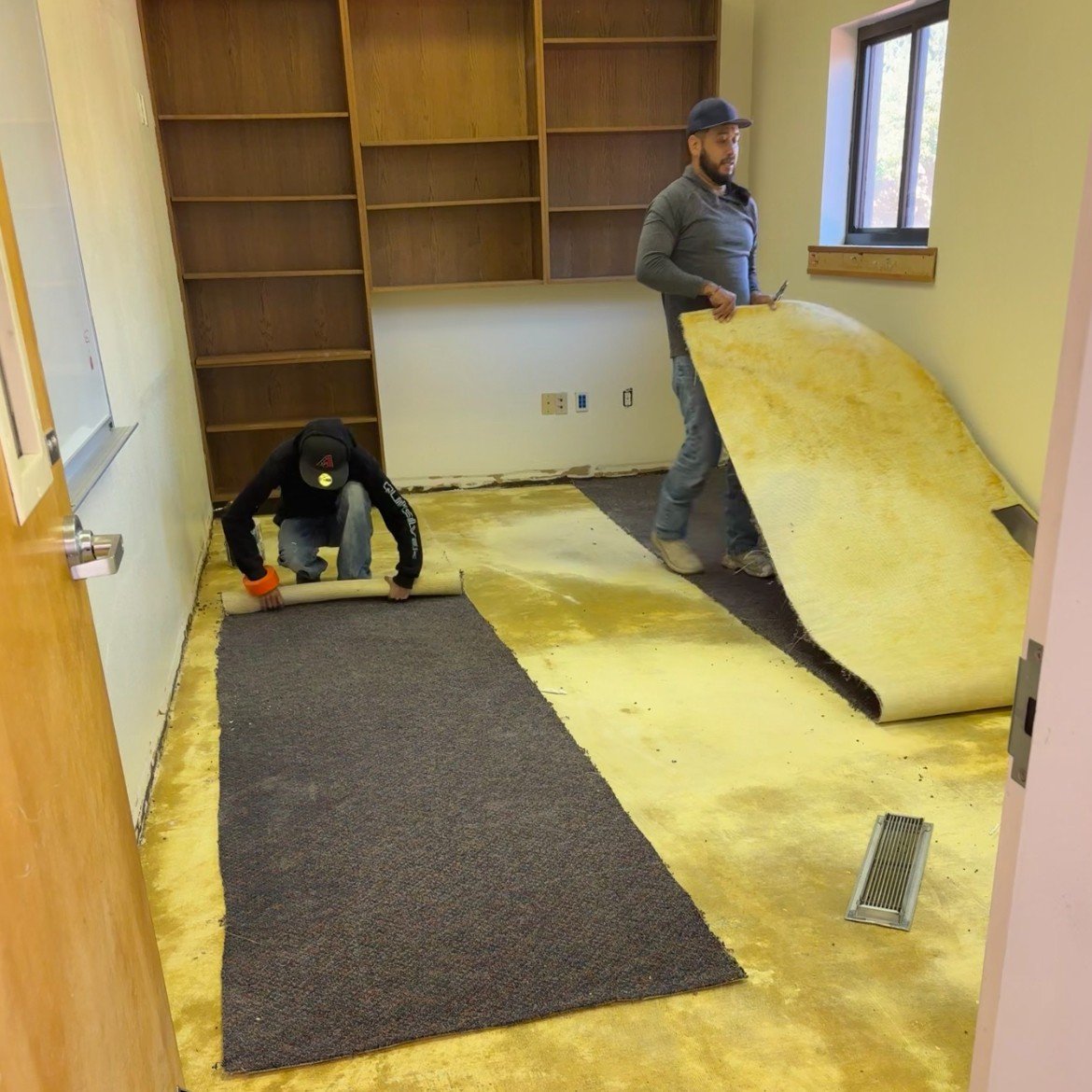 Two workers are removing old carpet flooring in a room with built-in wooden shelves, yellowish walls, and a window. One worker is rolling up the carpet near the doorway, and the other is lifting a section of carpet near the window.