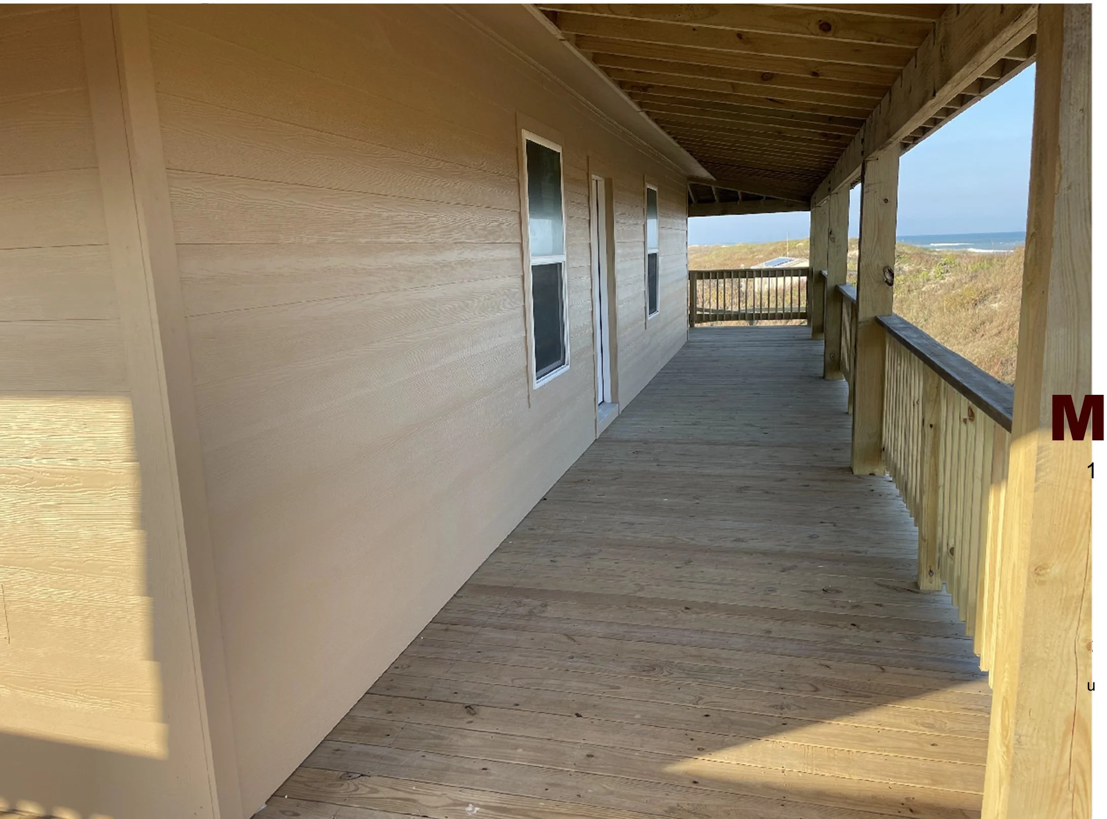 A wooden balcony with a railing overlooking a beach and ocean in the distance, under a clear blue sky.