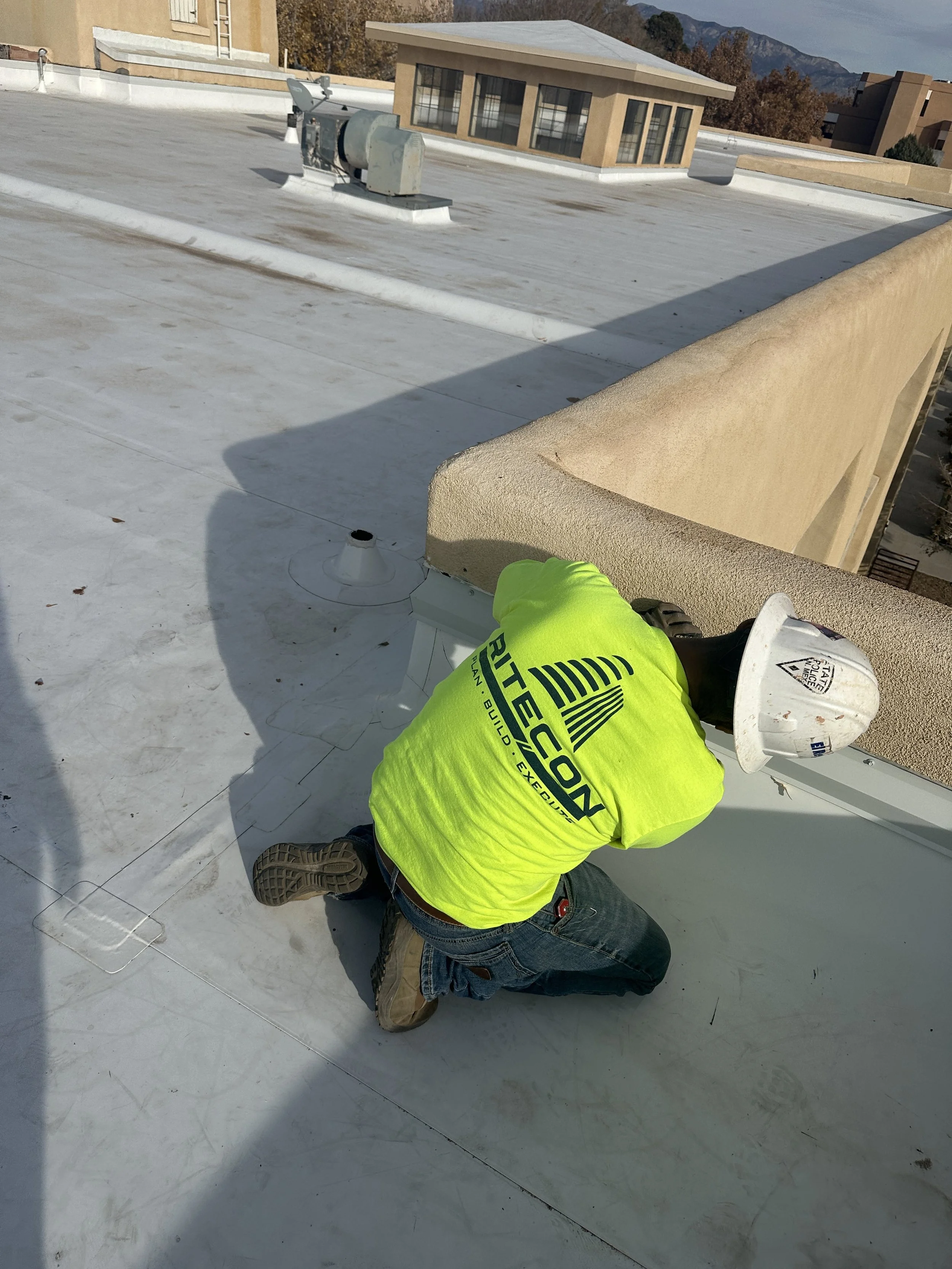 A construction worker wearing a bright yellow safety jacket and a white hard hat kneels on a flat rooftop, working on HVAC equipment. The rooftop has a white surface with some dirt and debris, and controlling equipment and vents are visible. In the b